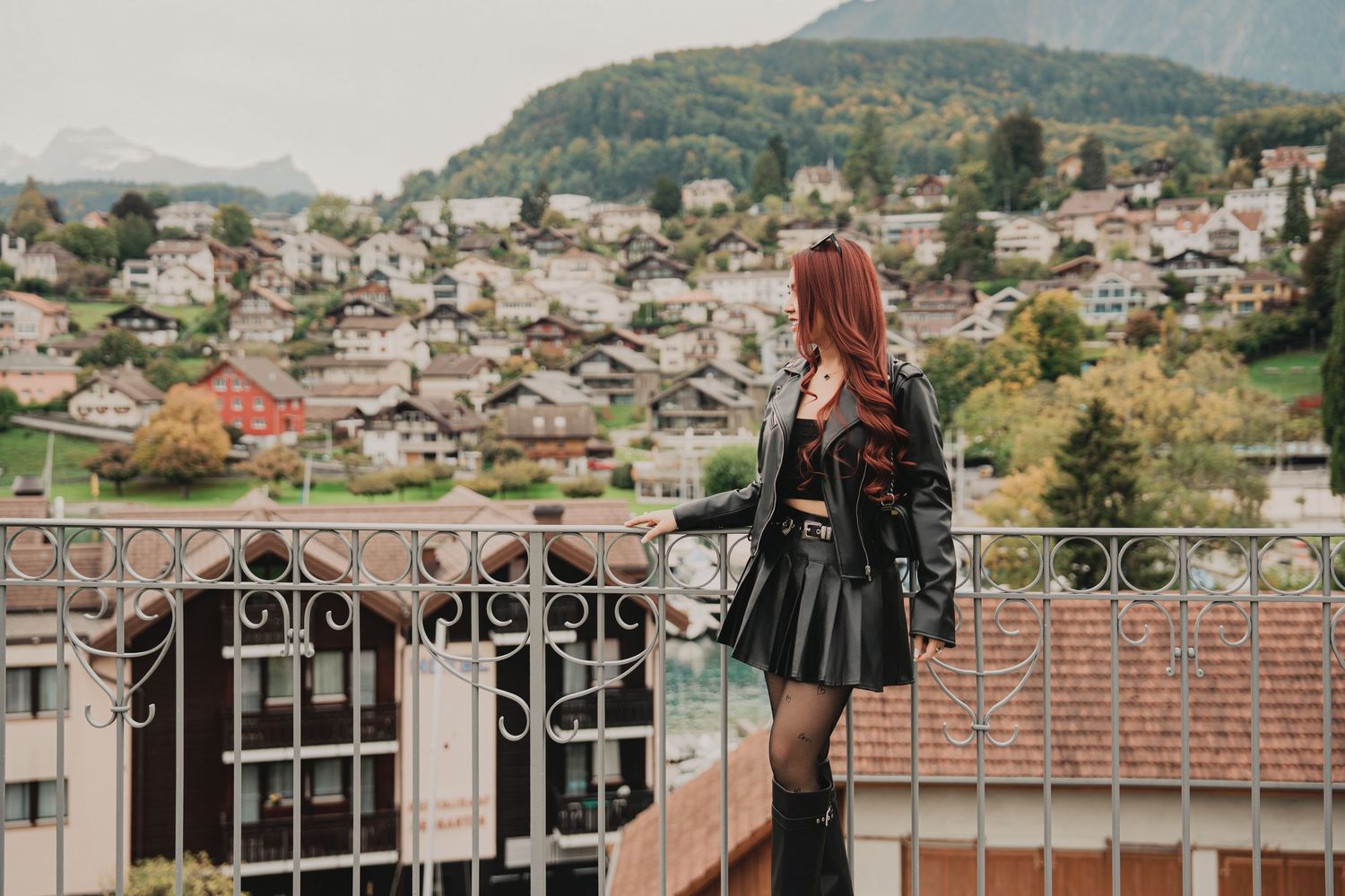 Stylish woman in black leather outfit standing on a balcony overlooking a European mountain town.