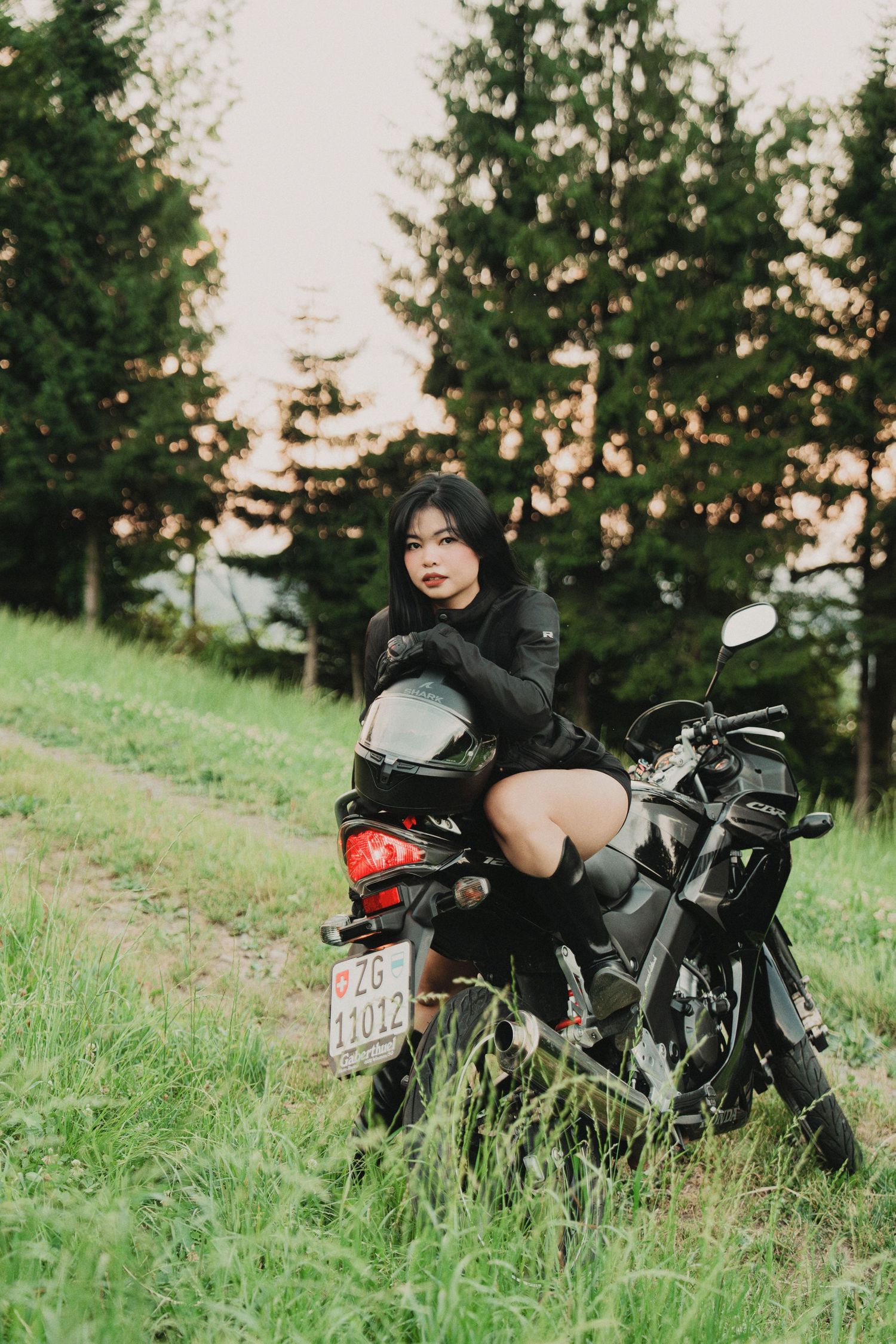 Young Asian woman sitting on black motorcycle on rural grass path with pine trees behind.