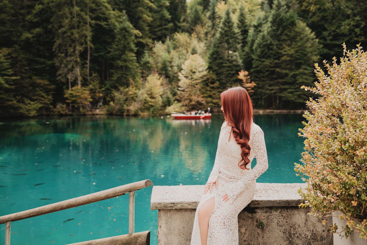 Red-haired woman in white lace dress sitting on stone railing overlooking turquoise mountain lake.
