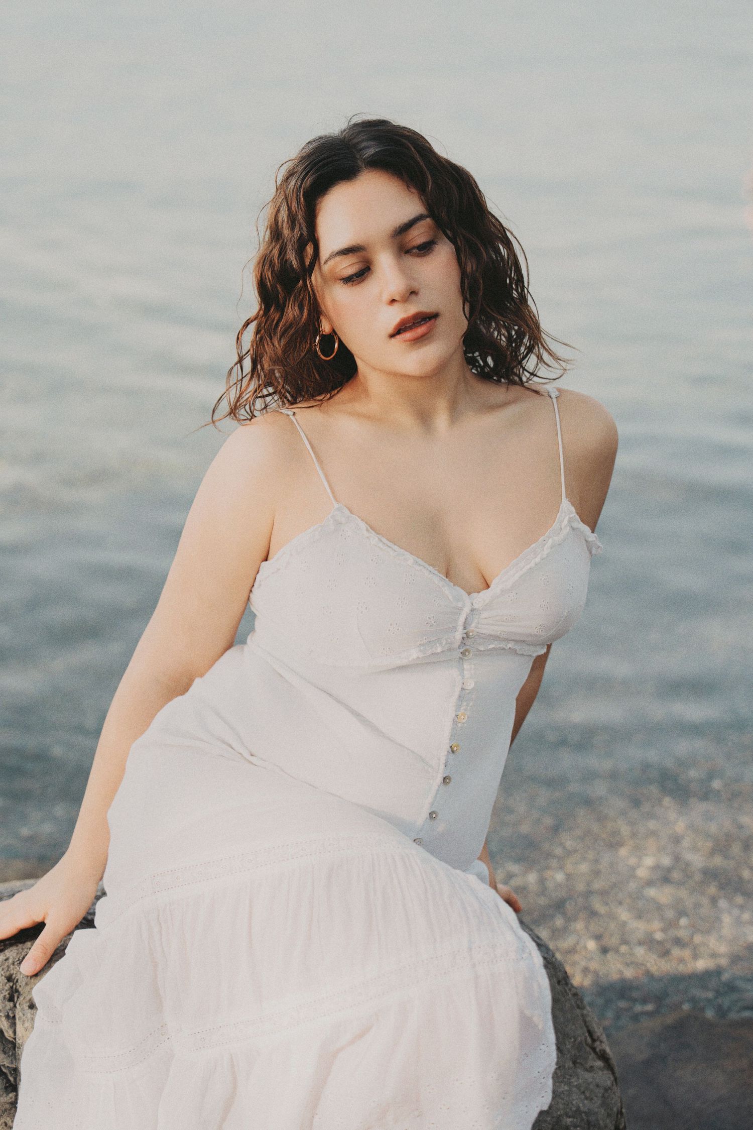 Young woman in white lace sundress sits on seaside rocks with eyes closed near calm water at dusk.