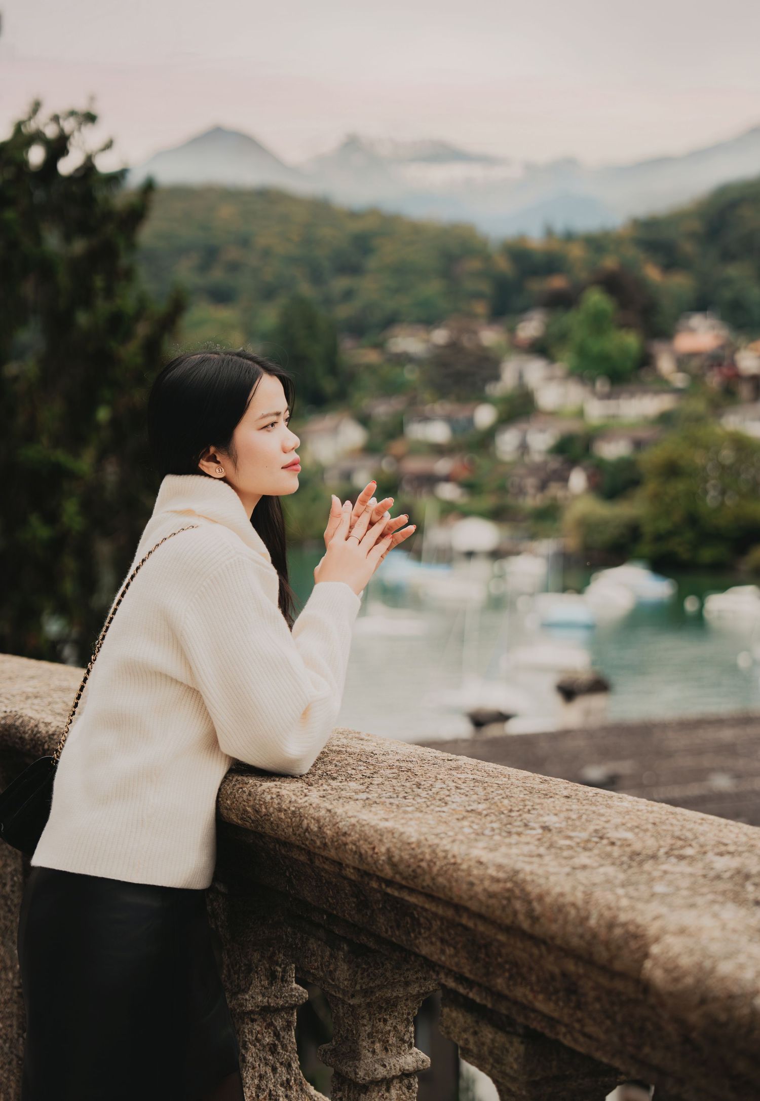 Woman in white sweater rests hands on stone balustrade overlooking scenic Swiss mountain lake village.