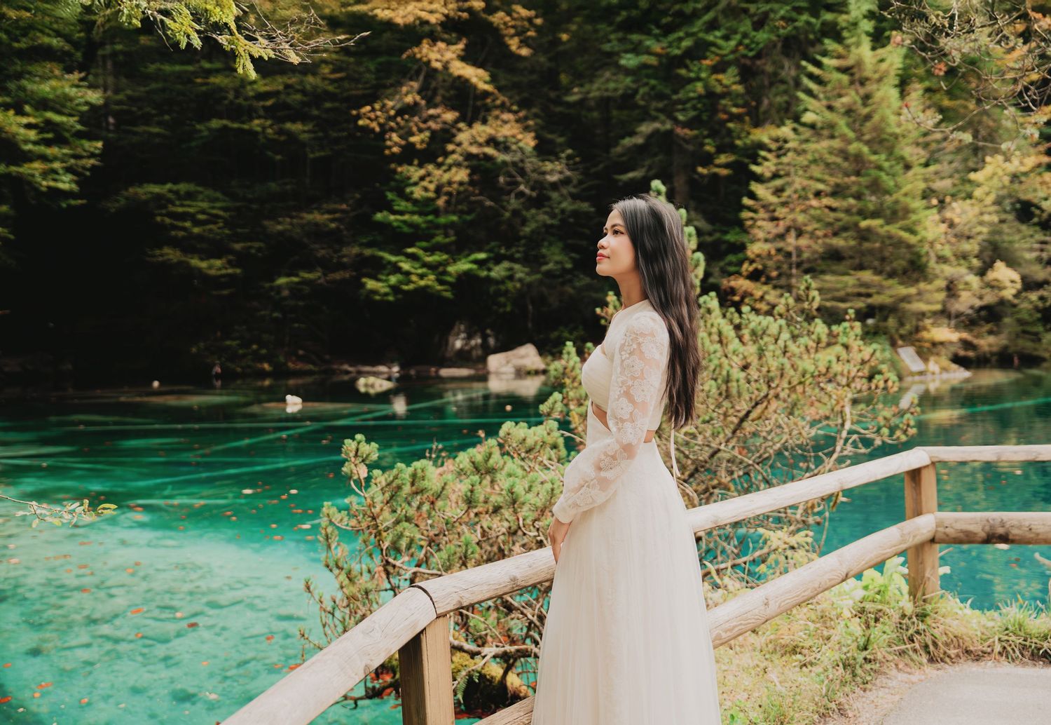 Young woman in white lace dress standing by wooden fence admiring a vivid turquoise lake in a forest.