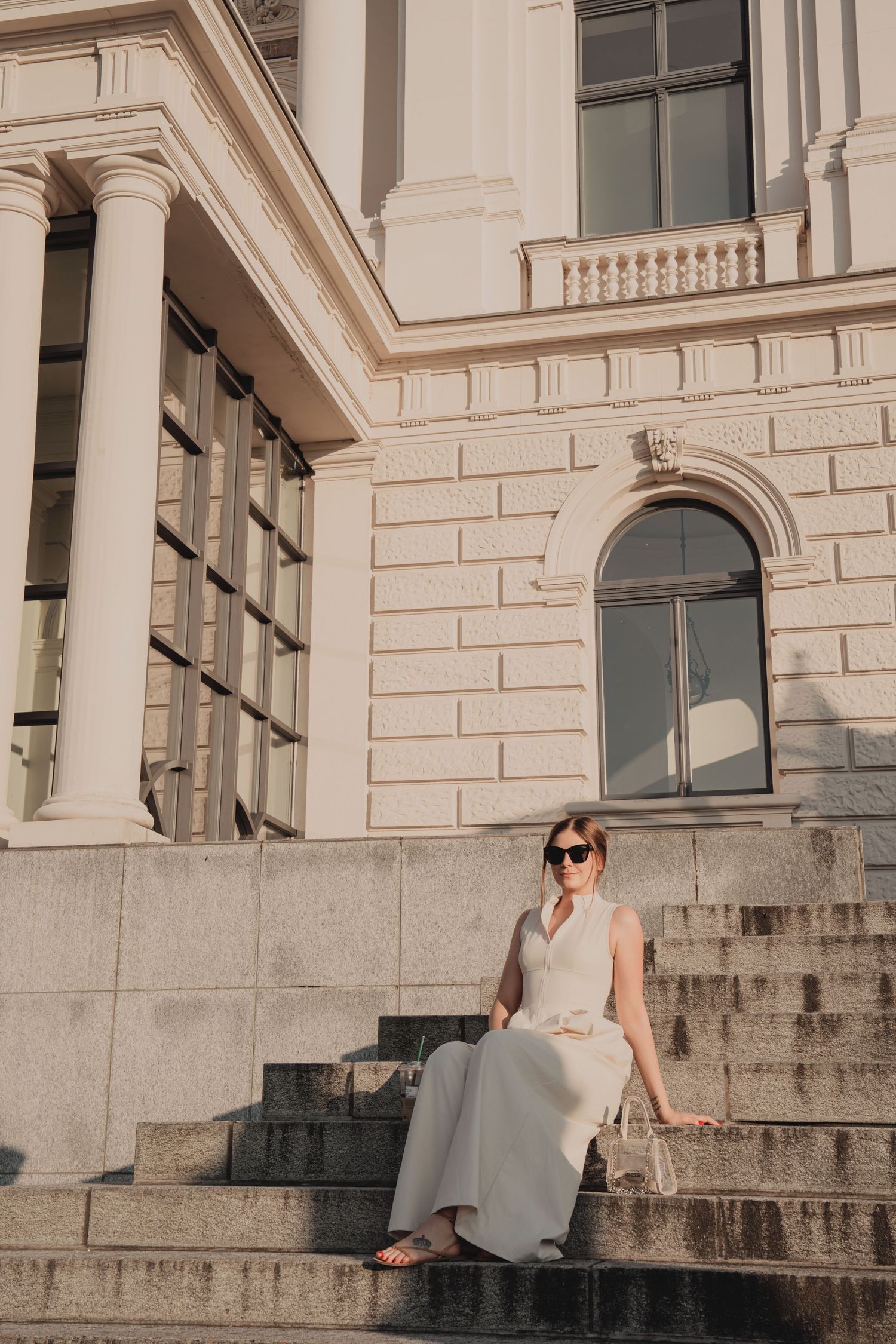 Elegant woman in white dress sitting on stone steps of classical European building.