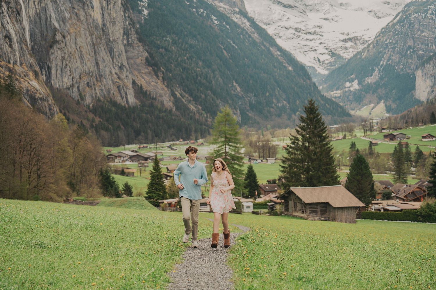 Couple walking hand in hand on a path through a lush green valley with Swiss Alps and charming village in background.