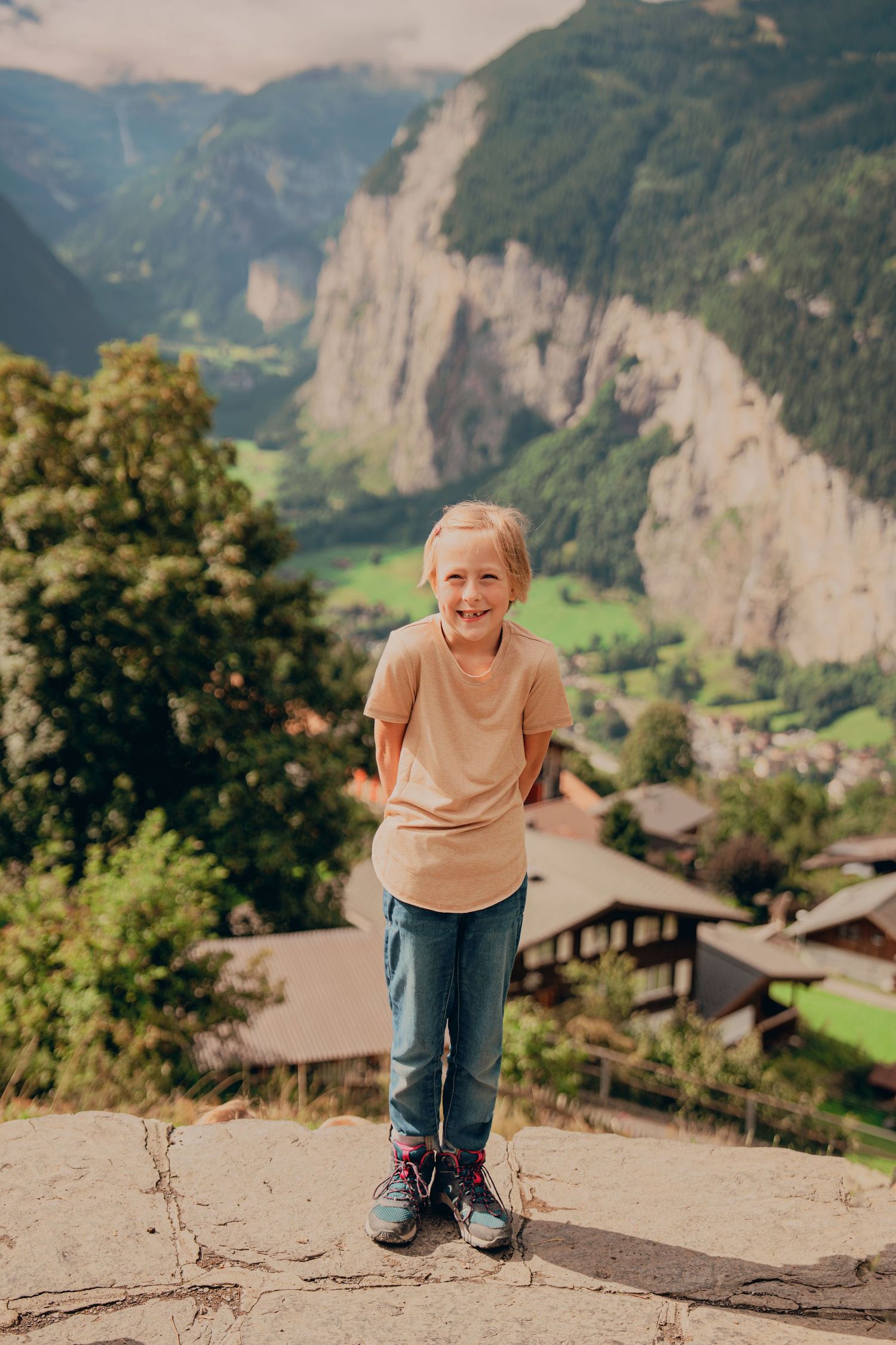 Laughing blonde child in beige shirt and jeans posing on stone ledge with Swiss mountain village below.