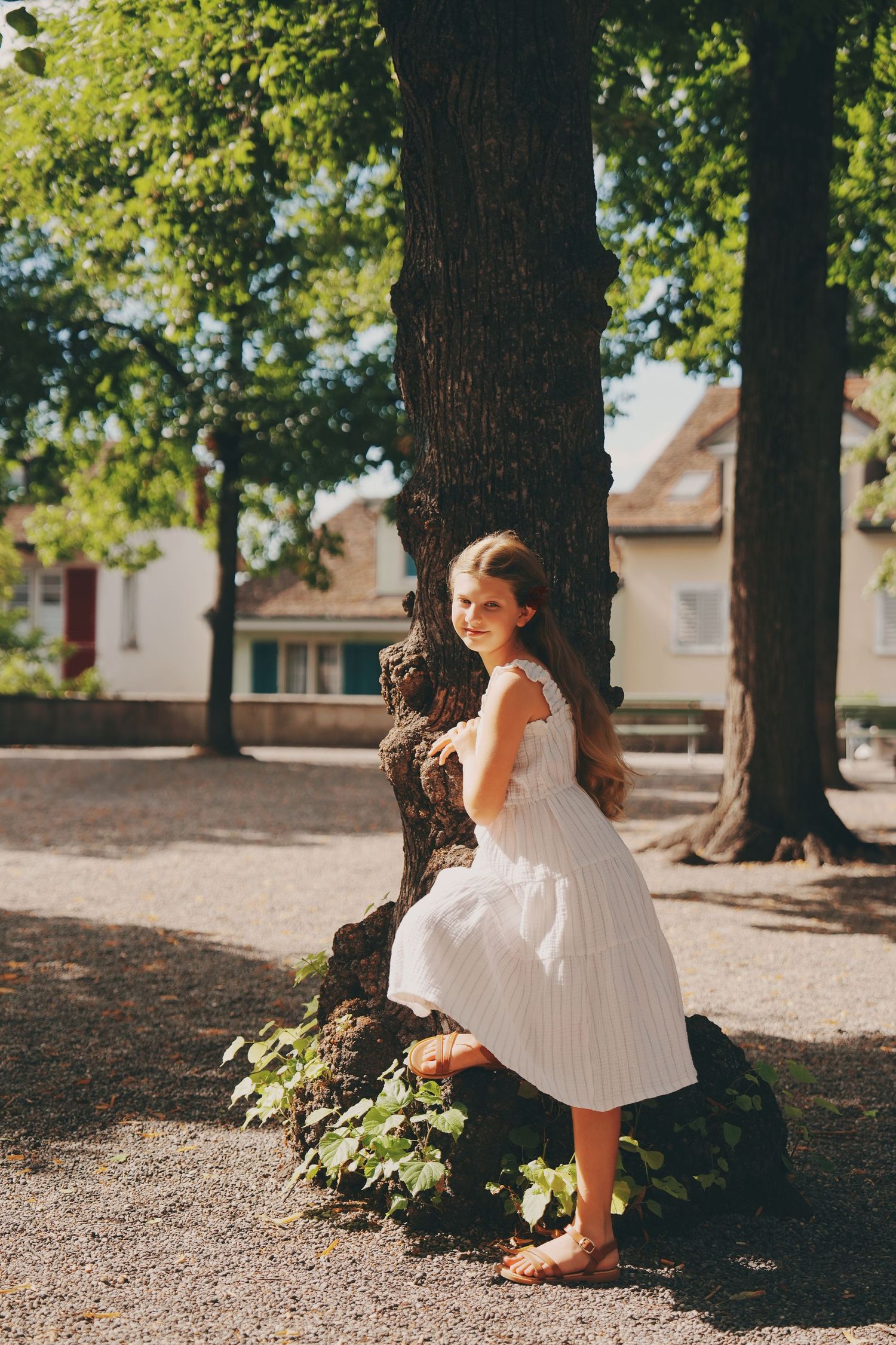 A young girl in a white striped dress leans against a large tree trunk in a sunny park.
