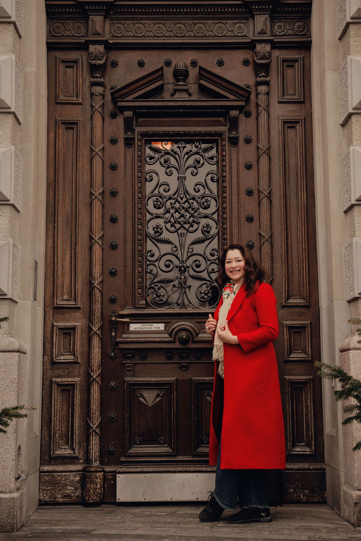 Woman in elegant red coat standing alone before a grand ornate carved wooden door.