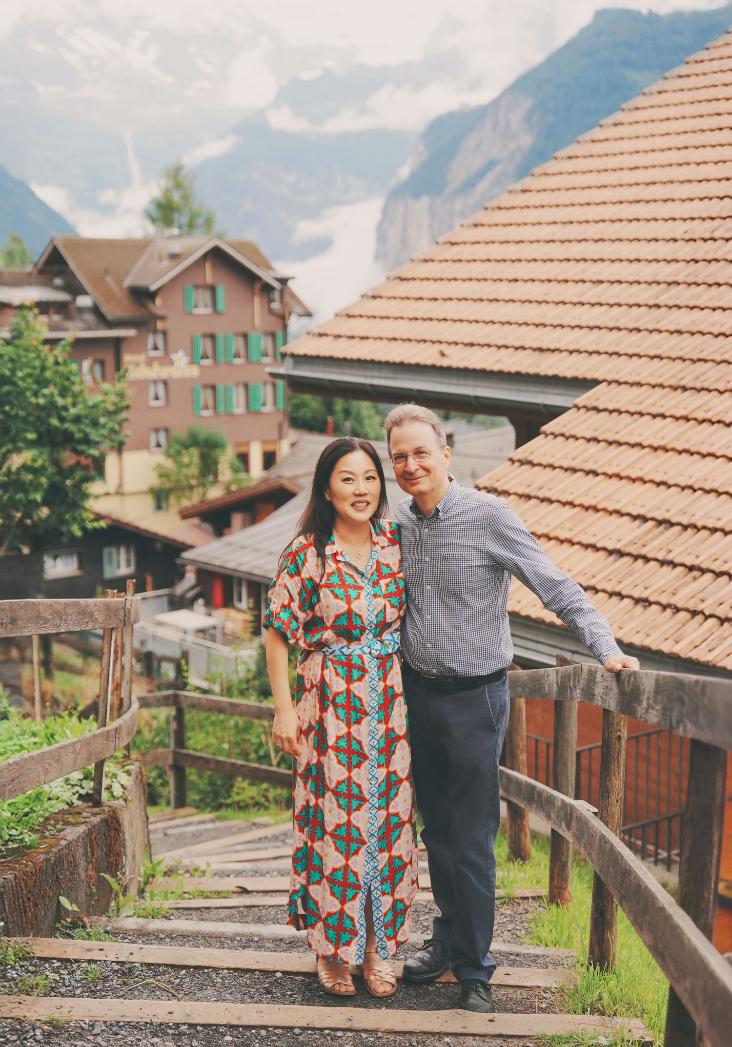 Smiling couple stands on stone steps before colorful Swiss village buildings and snow-capped Alpine peaks.