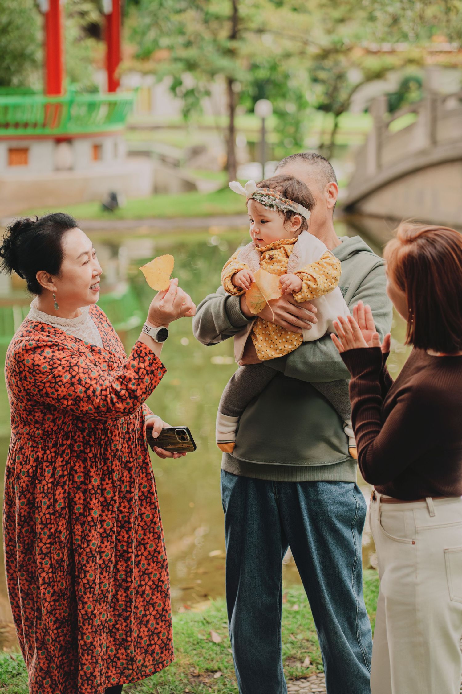 Asian family giving red envelopes to baby near scenic pond with arched stone bridge.