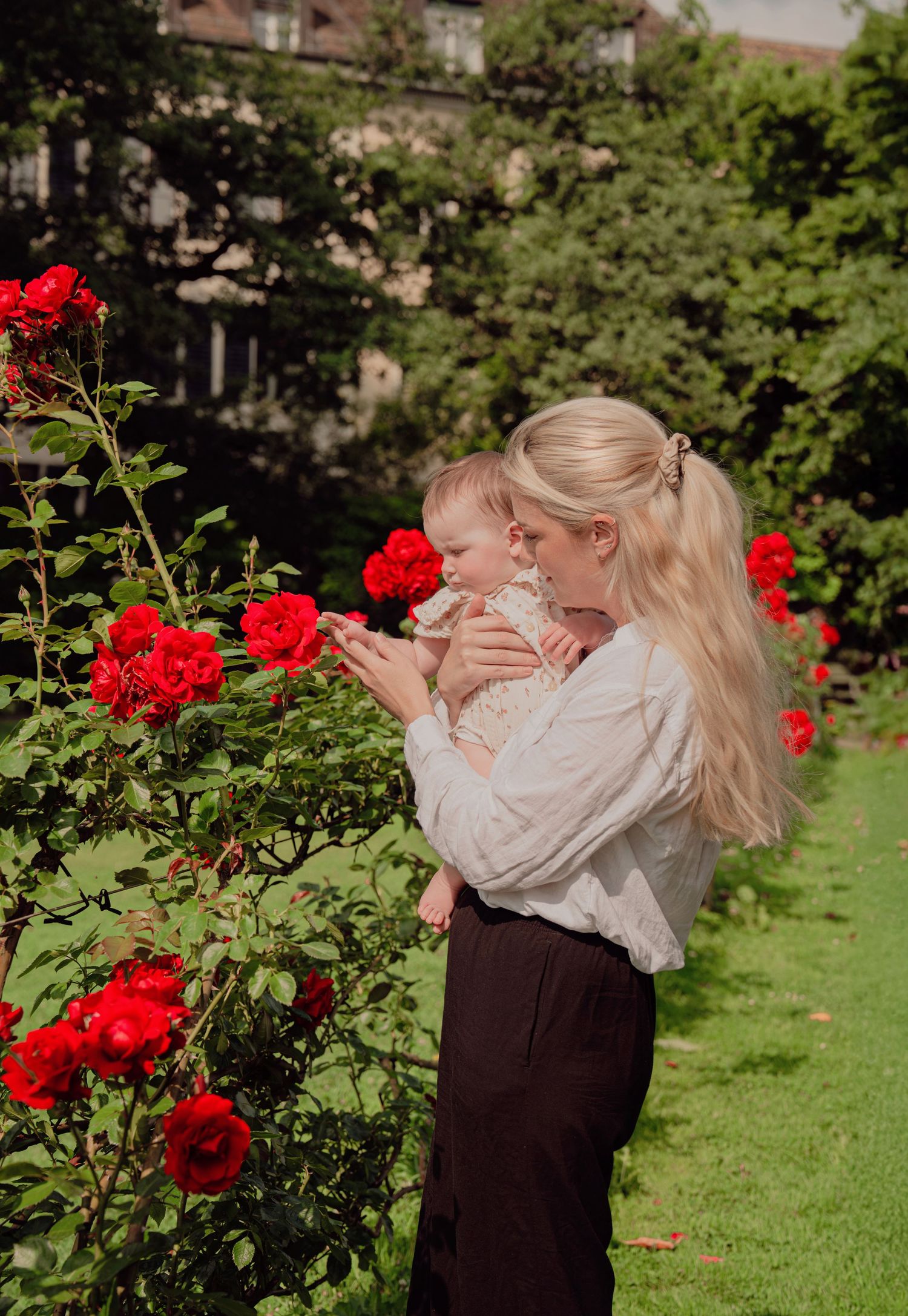 Blonde mother holding baby near vibrant red roses in a green garden.