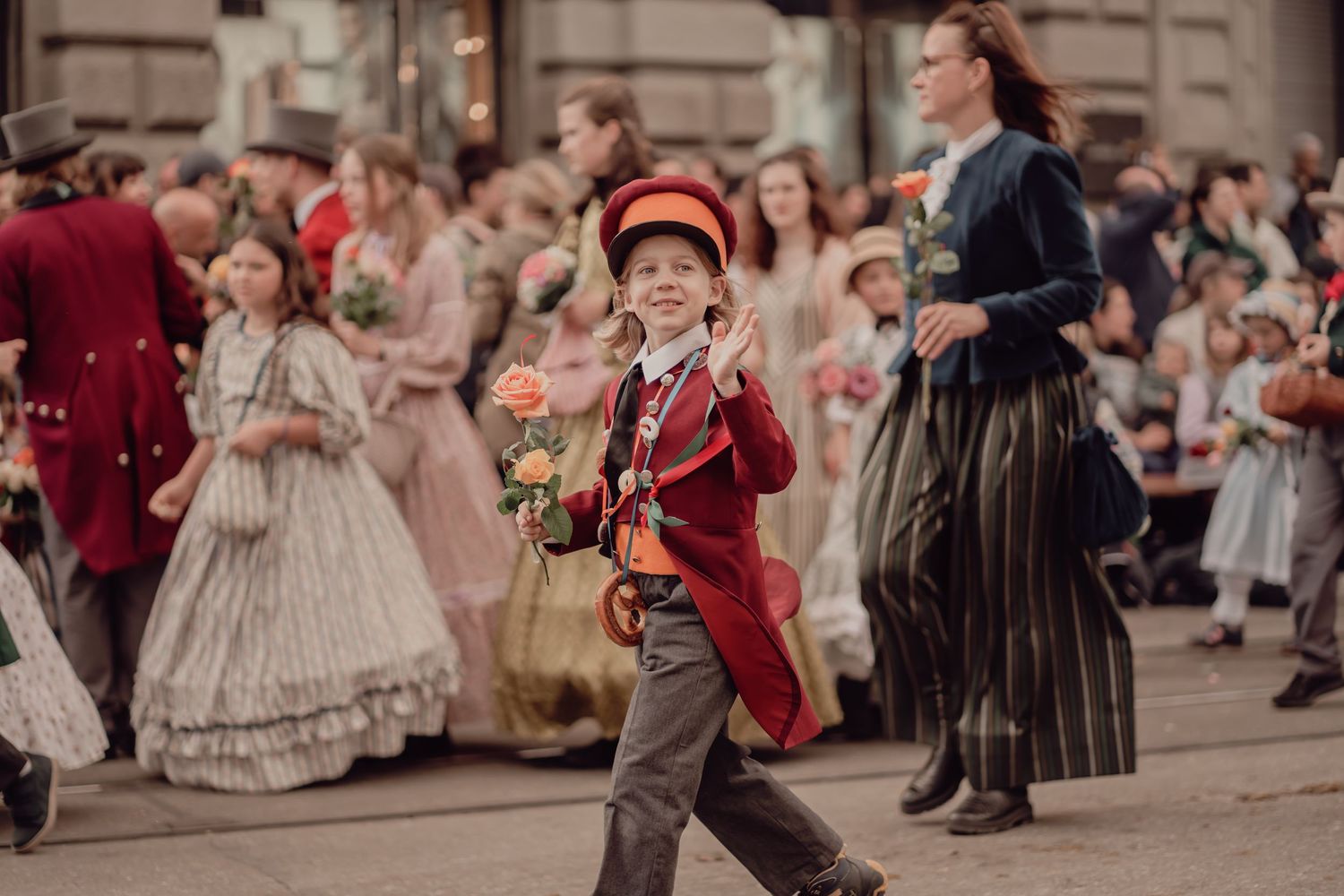 Cheerful boy in red vintage uniform marches with roses during a festive historical street parade.