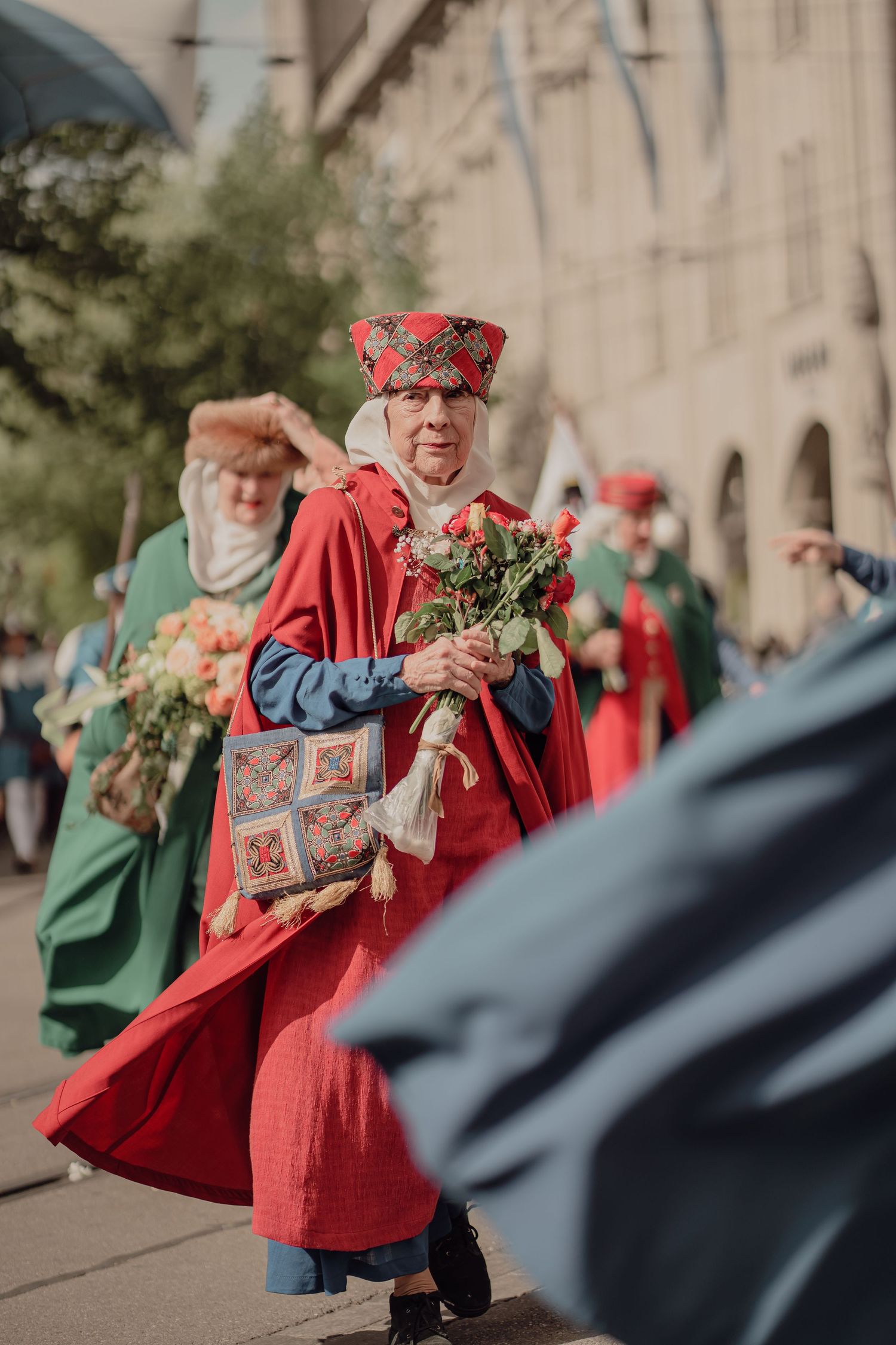 Man in elaborate red medieval costume with ornate hat carries roses during historical cultural parade.