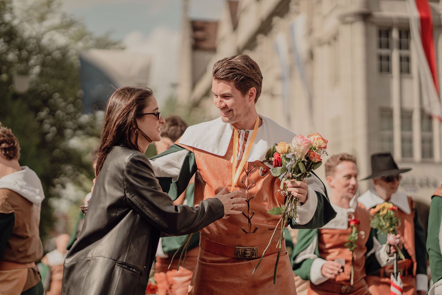 A woman exchanges a medal with a young man in traditional orange costume during a historic street festival.