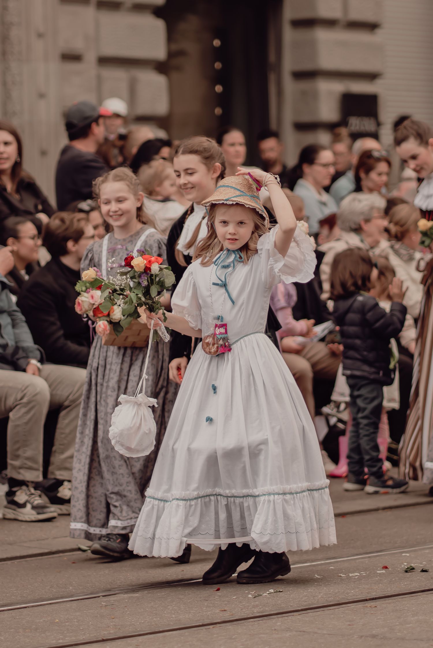 A little girl in a white vintage dress adjusts her straw hat while carrying flowers in a parade.