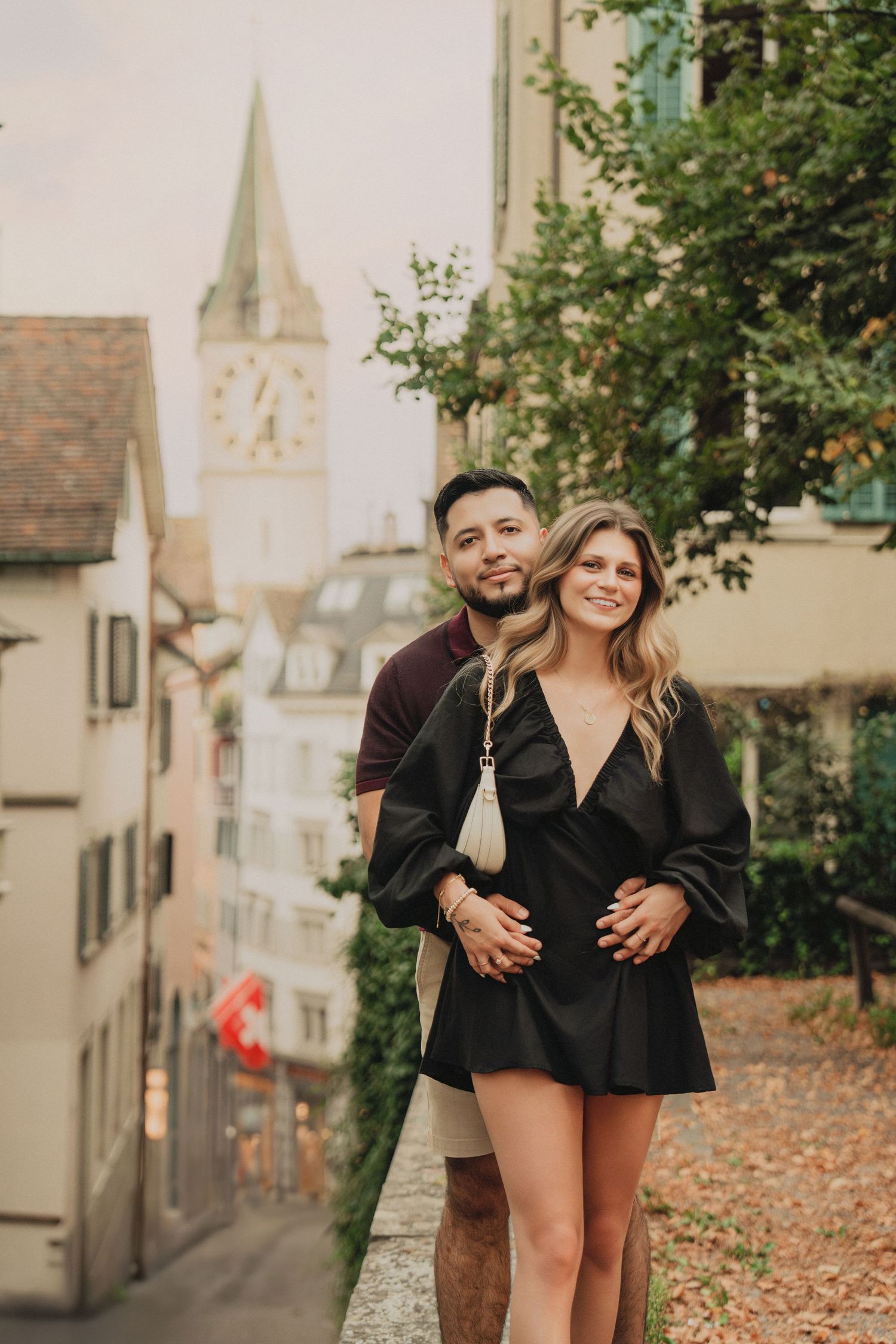 Couple embracing on a scenic Zurich alley with St. Peter's Church tower visible in the soft background.