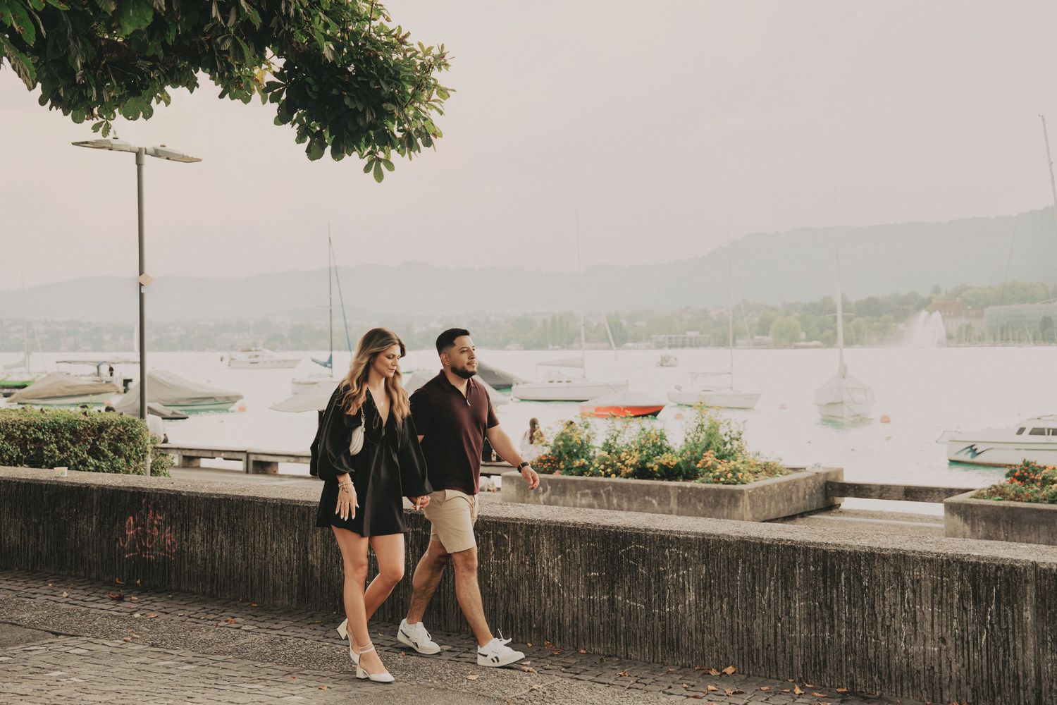 Couple walking along a lakeside promenade in Zurich with sailboats and mountains in the background.