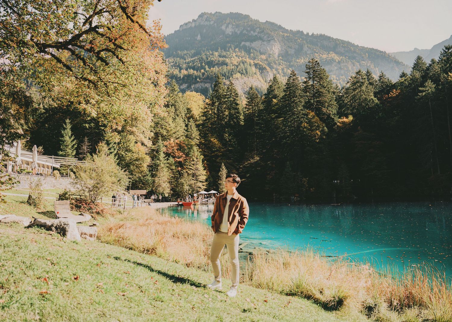 Man standing near a brilliant turquoise alpine lake with autumn trees and mountain scenery.