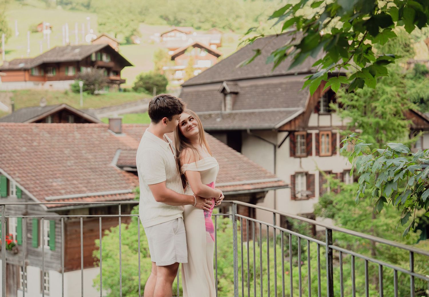 Young couple embracing on balcony surrounded by traditional Swiss chalet houses and green hills.