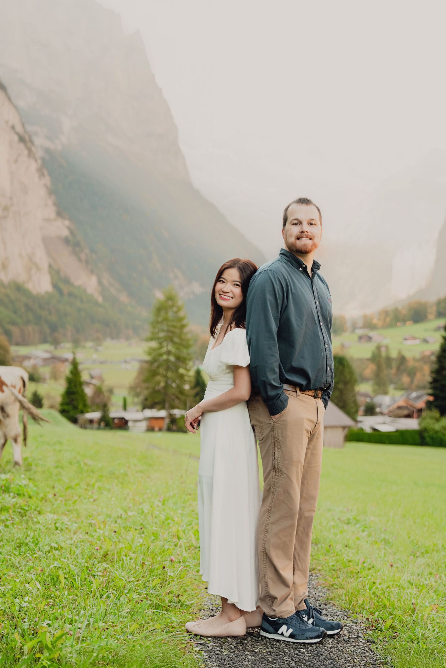 Smiling couple standing together on a scenic path with a misty Swiss mountain valley and green meadows behind them.
