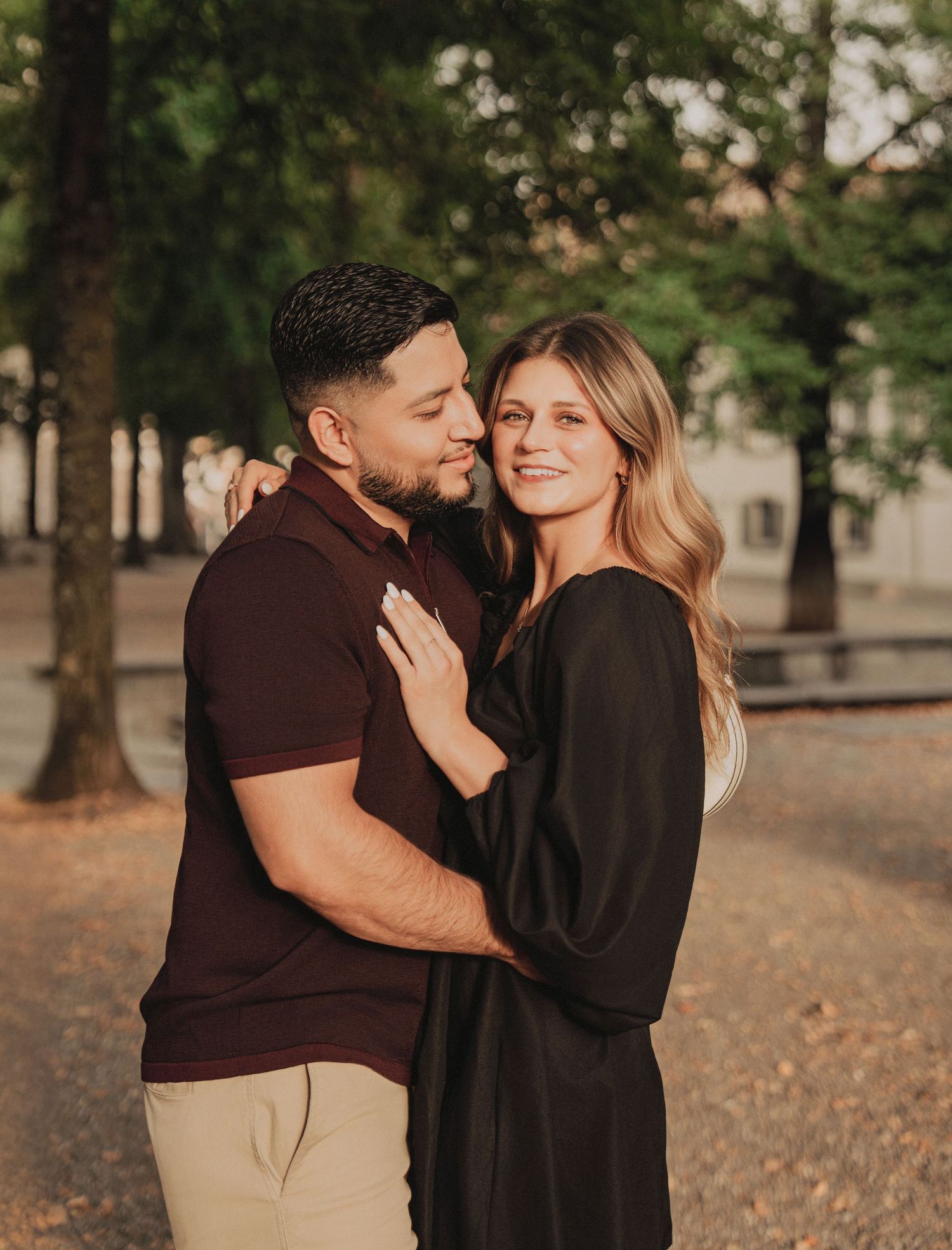 Smiling couple embracing warmly in a tree-lined park with golden hour light in a European city.