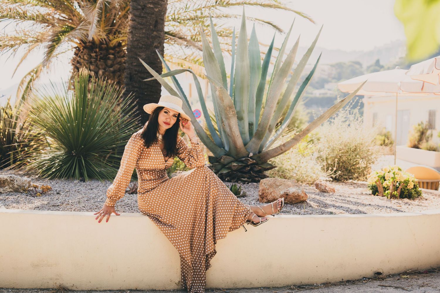 Woman in polka dot maxi dress and sun hat sitting by desert garden with palm and agave plants.