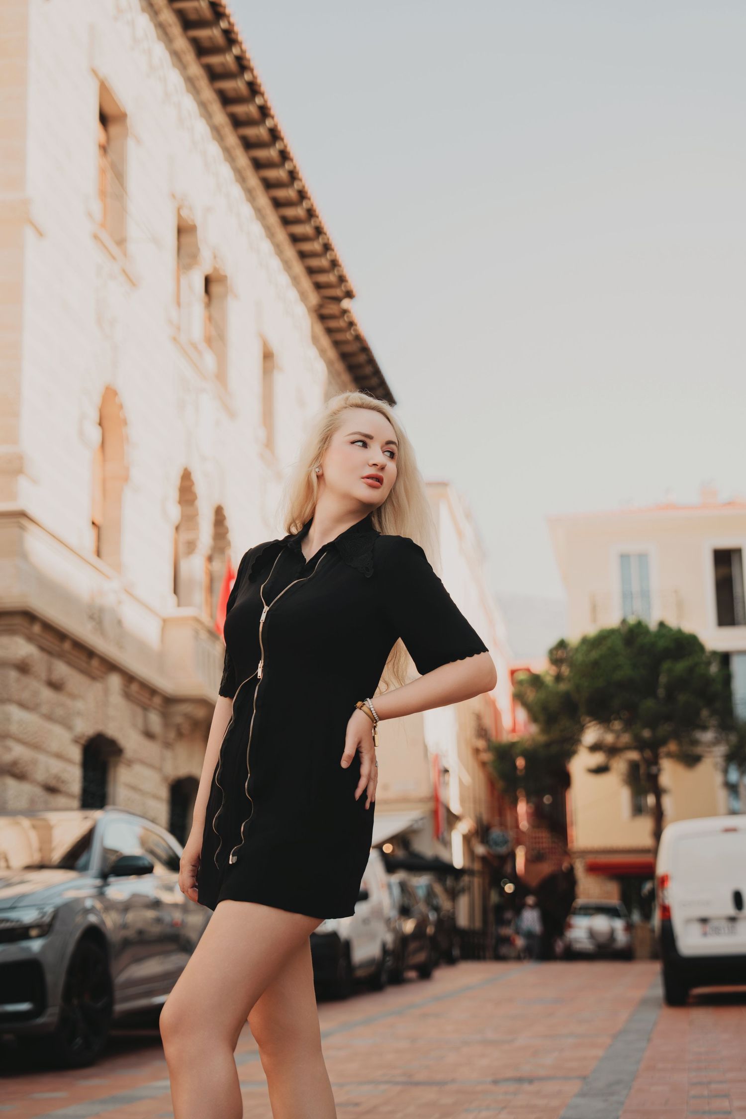 Blonde woman in black mini dress posing on a European city street with historic buildings.