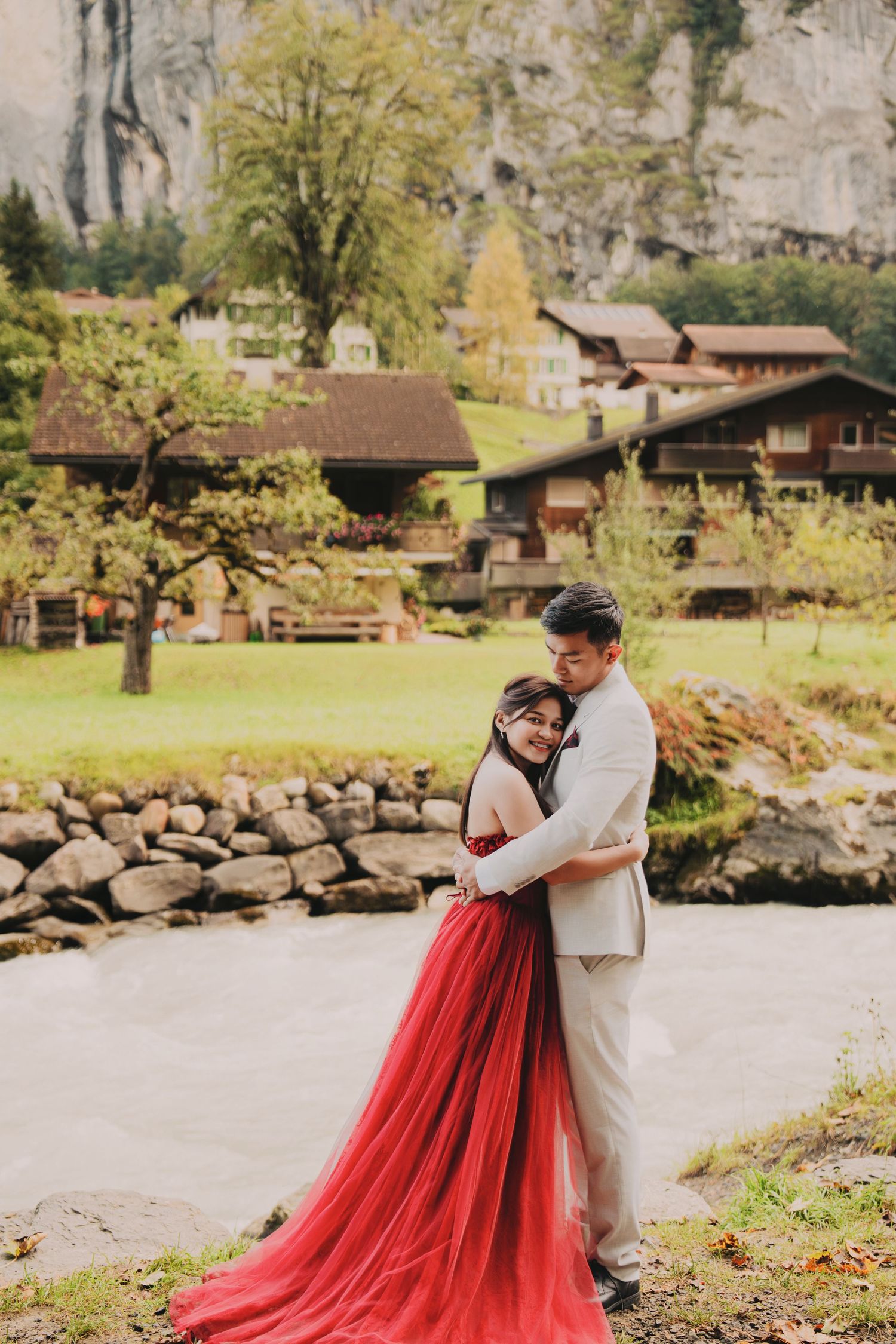 Romantic couple hugging near a rocky riverbank in a Swiss alpine village, bride wearing a flowing red dress.