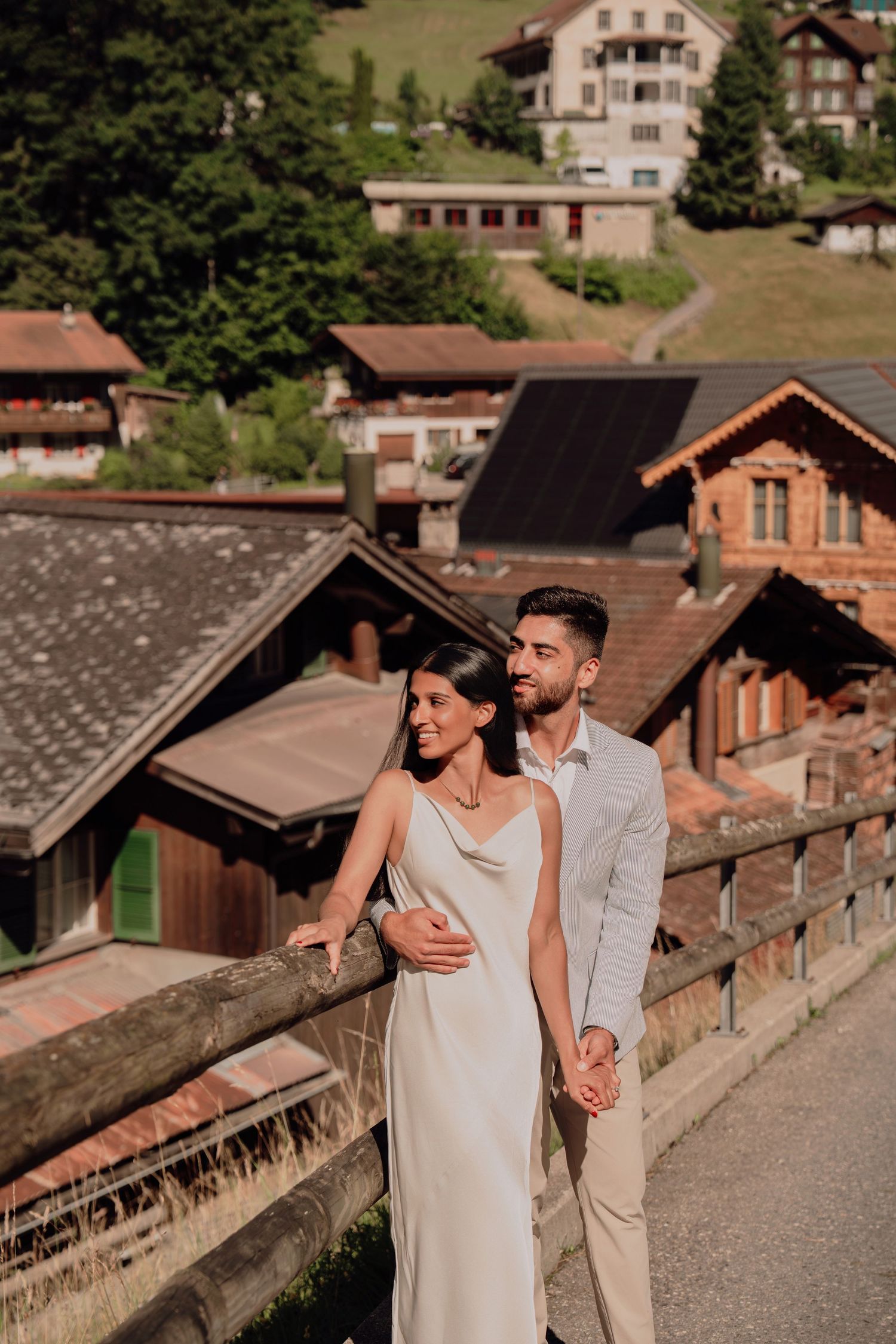 Elegant couple embraces on a bridge with rustic Alpine chalets and green hillside in the background.