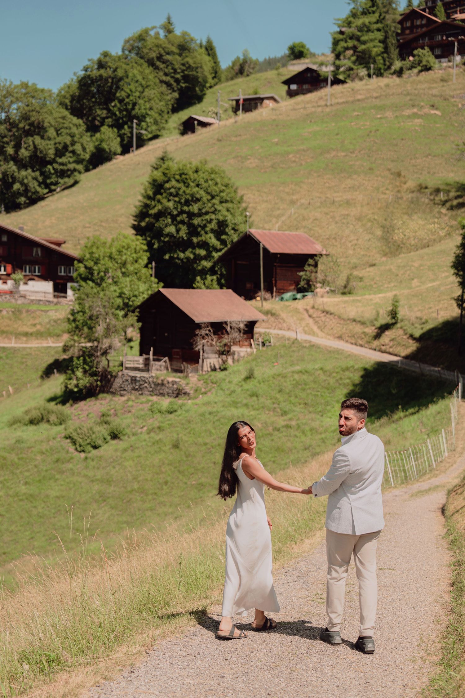 Wedding couple holding hands walking on a rural mountain path with green Swiss hillside and chalets.