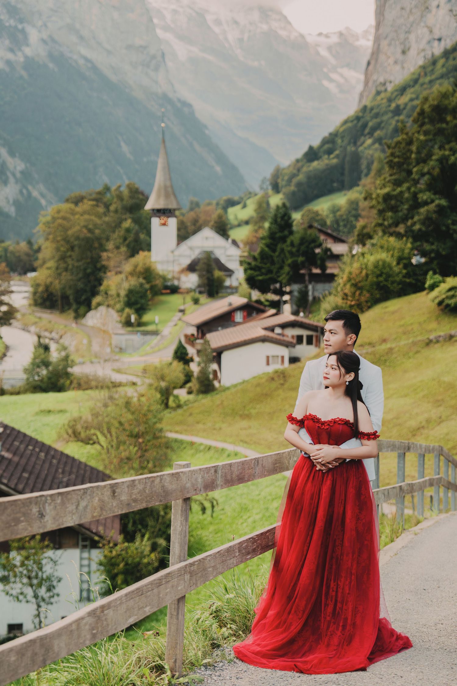 Couple in red gown and suit embracing on a bridge with Swiss alpine village and mountains backdrop.