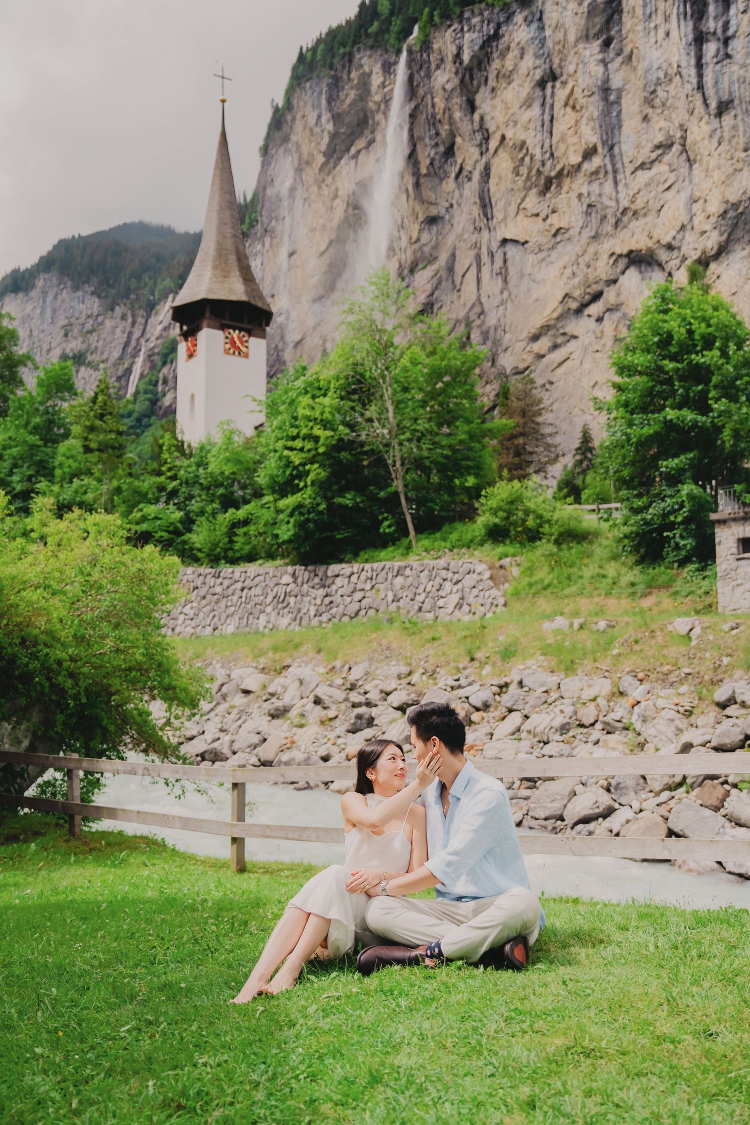 Couple sitting on grass in Lauterbrunnen valley with church tower and waterfall backdrop in Swiss Alps.
