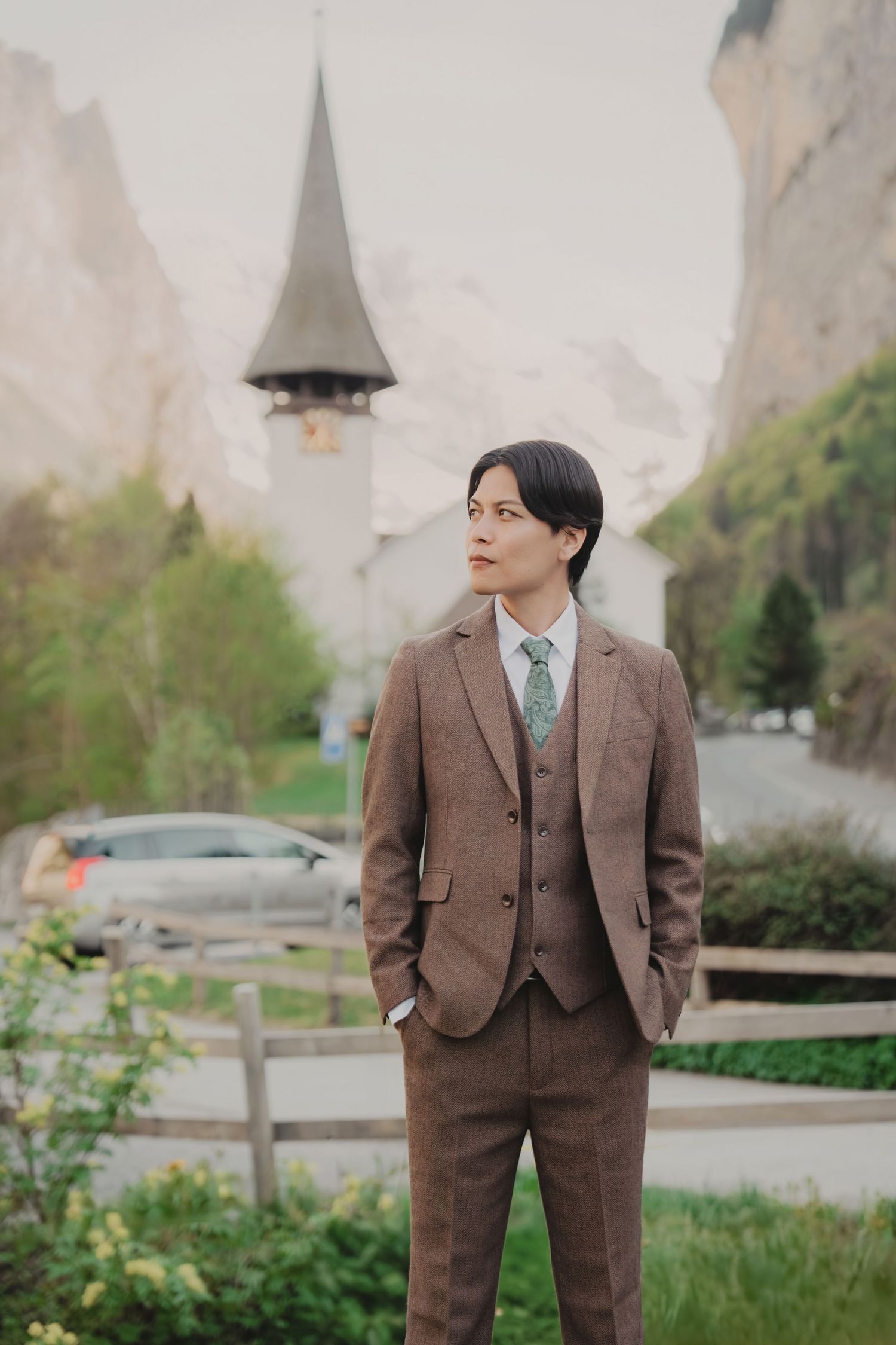 Groom in brown three-piece suit stands confidently before a Swiss alpine church tower.