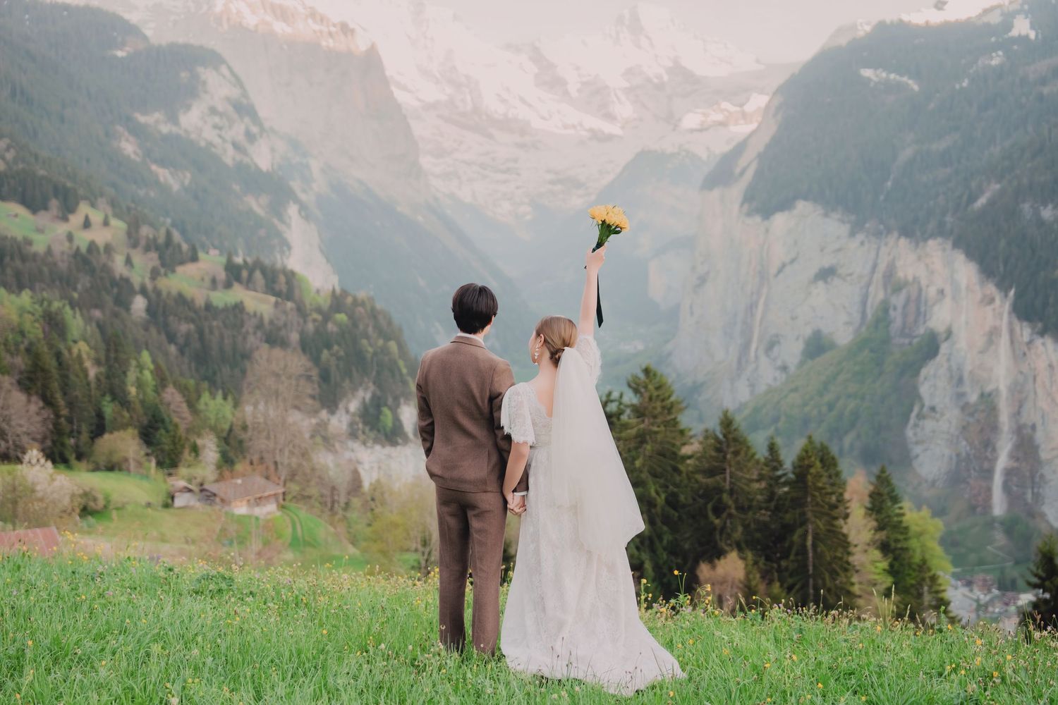 Bride and groom raising bouquet triumphantly while facing majestic Swiss Alps mountain landscape.