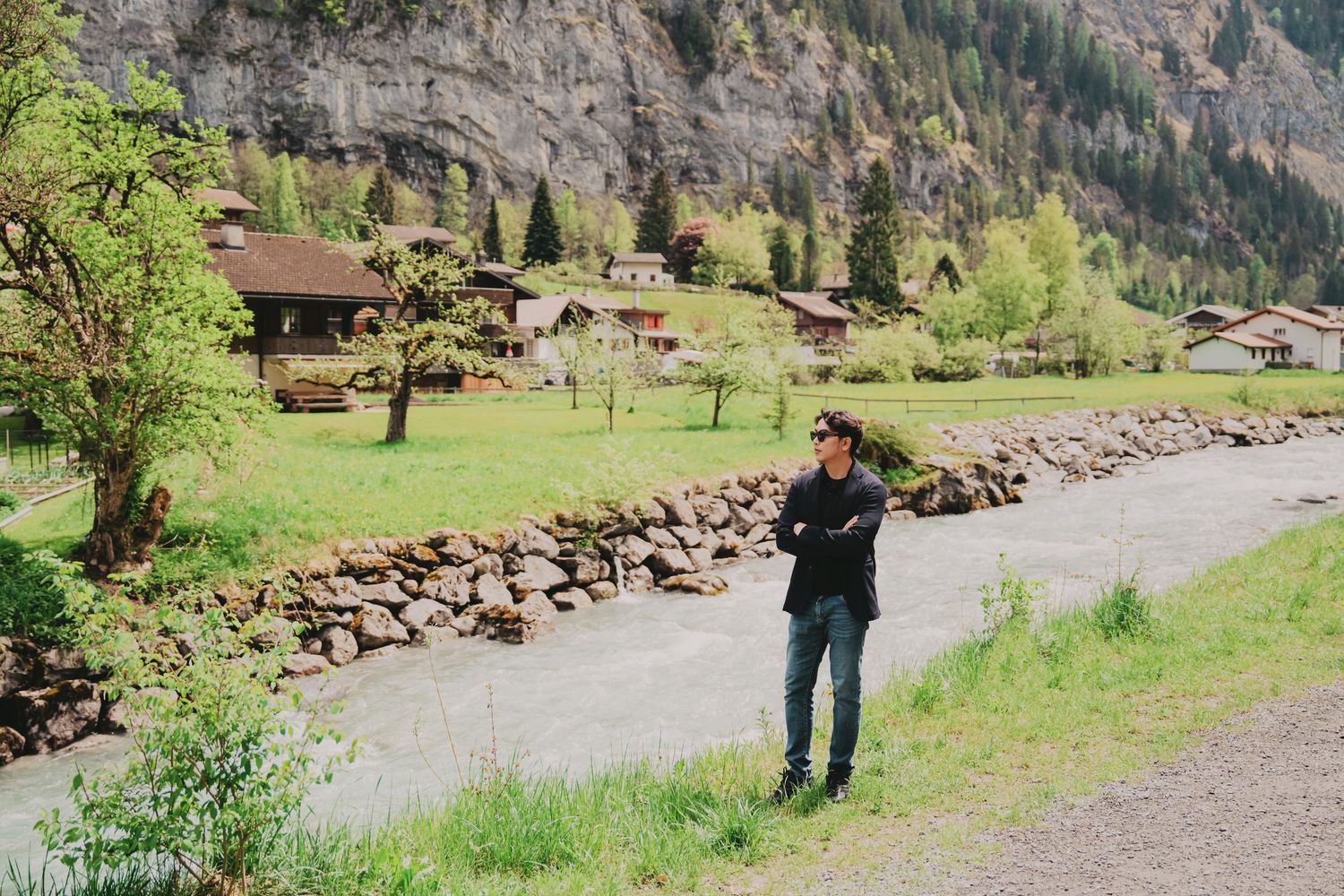 Man in black shirt standing on path beside rocky wall with Swiss alpine village in background.