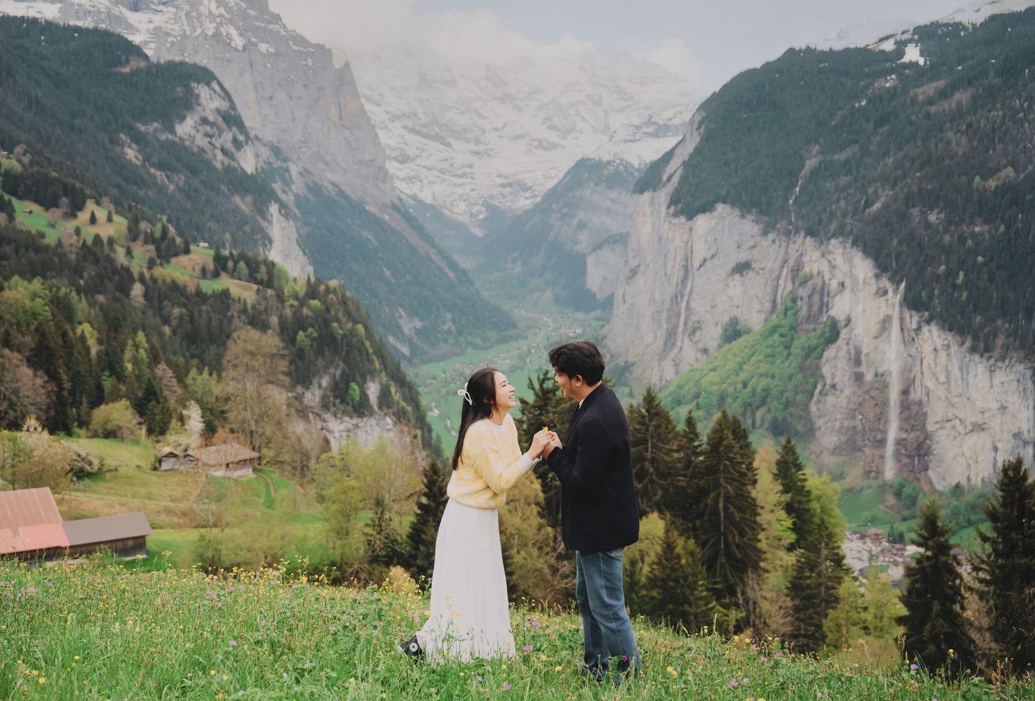 Couple standing together in Swiss alpine meadow with stunning snow-capped mountain valley behind them.