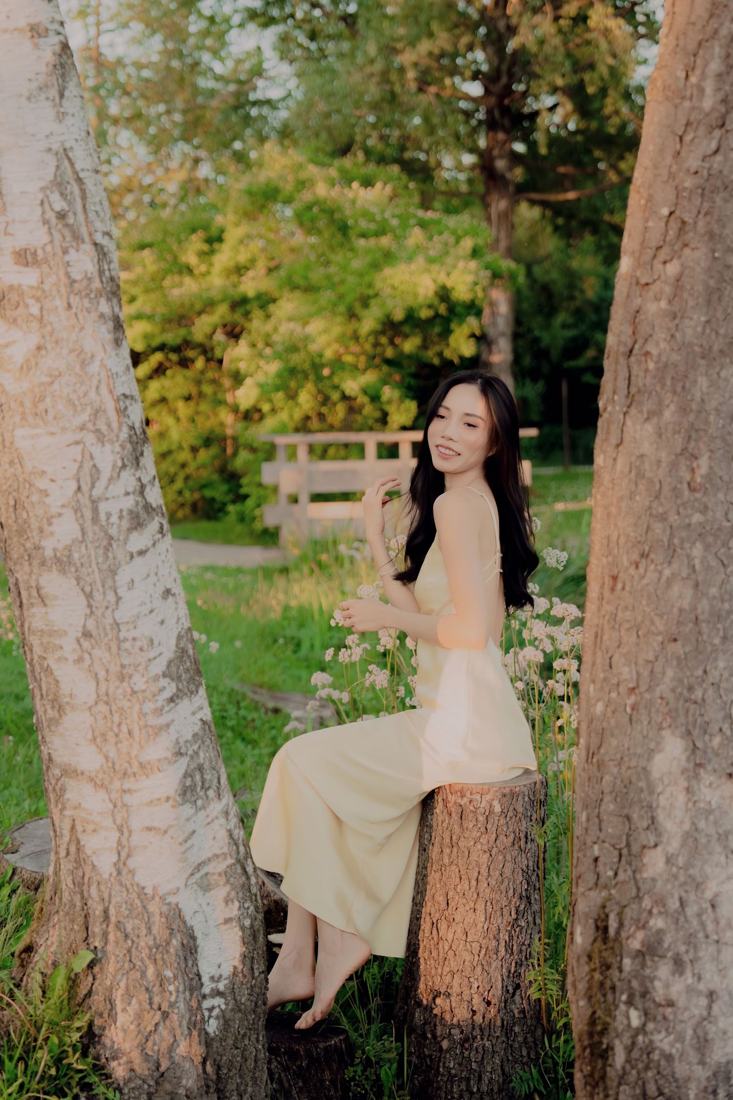 Young Asian woman in yellow dress sitting between birch trees in a sunlit garden smiling softly.