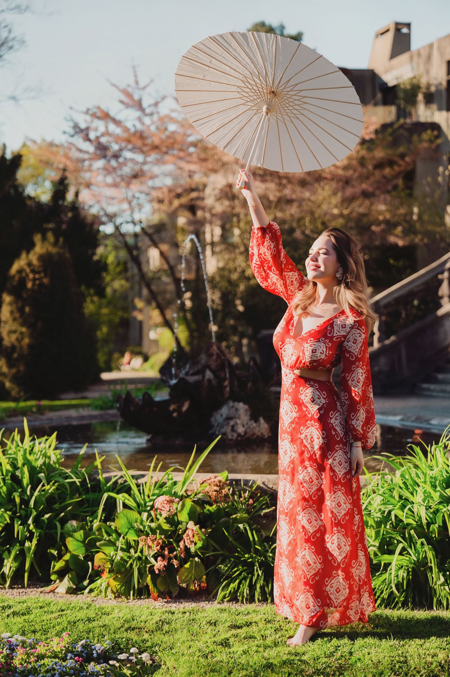 Smiling woman in red floral maxi dress raising white parasol in lush garden with fountain.