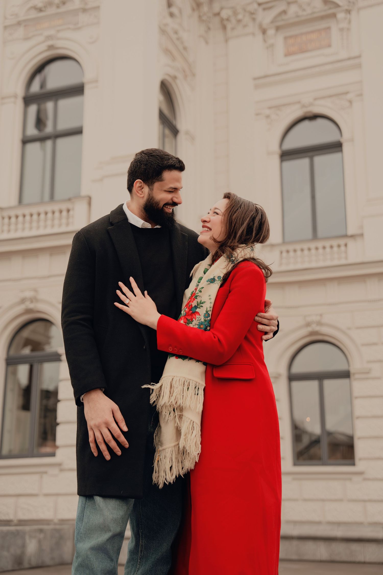 Smiling couple embraces in front of a white classical building, woman in red coat with floral scarf.