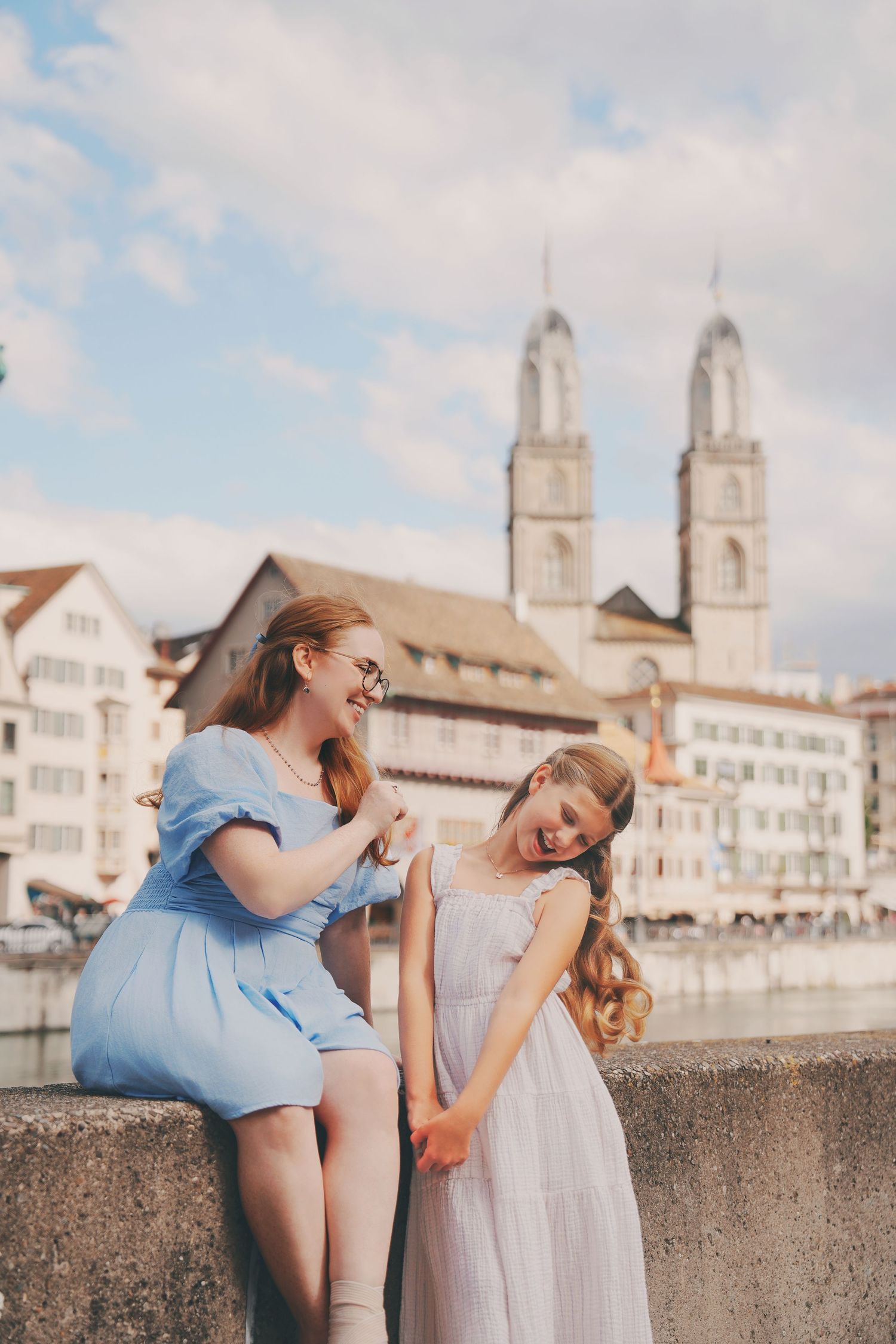 Redhead mother in blue dress and daughter in white laugh together on Zurich bridge.