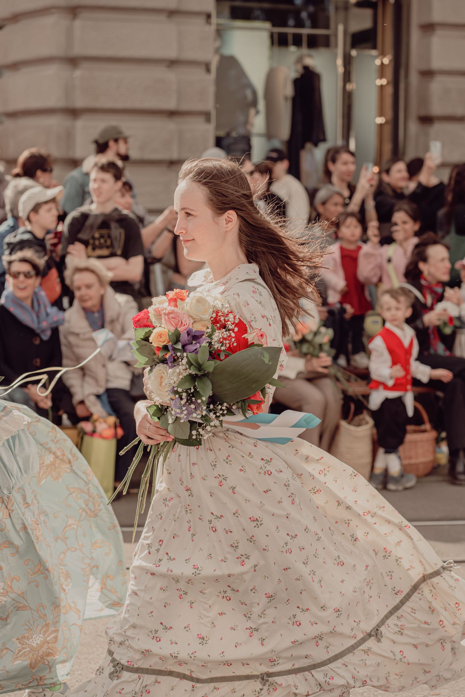 A woman in a floral vintage dress twirls with a bouquet of flowers during a festive street parade.