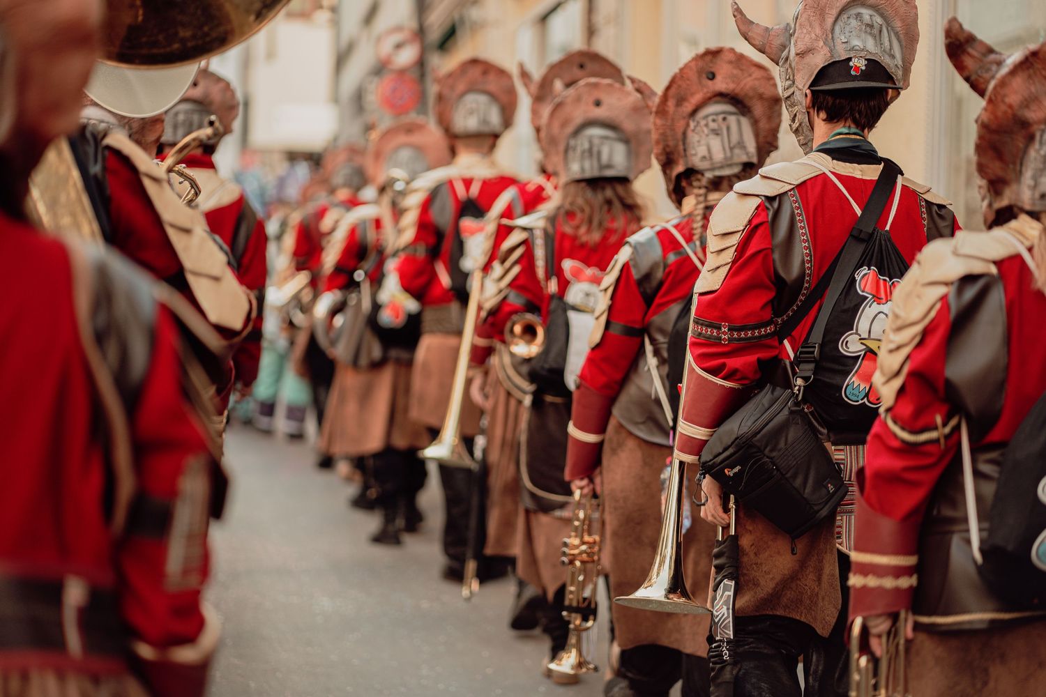 Marching band members in red historical military uniforms parade through a cobblestone street.