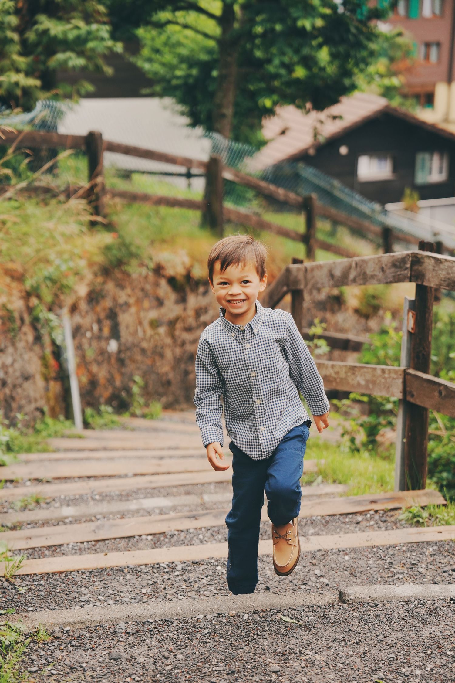 Cheerful young boy in plaid shirt runs up wooden outdoor stairs in a lush alpine village setting.