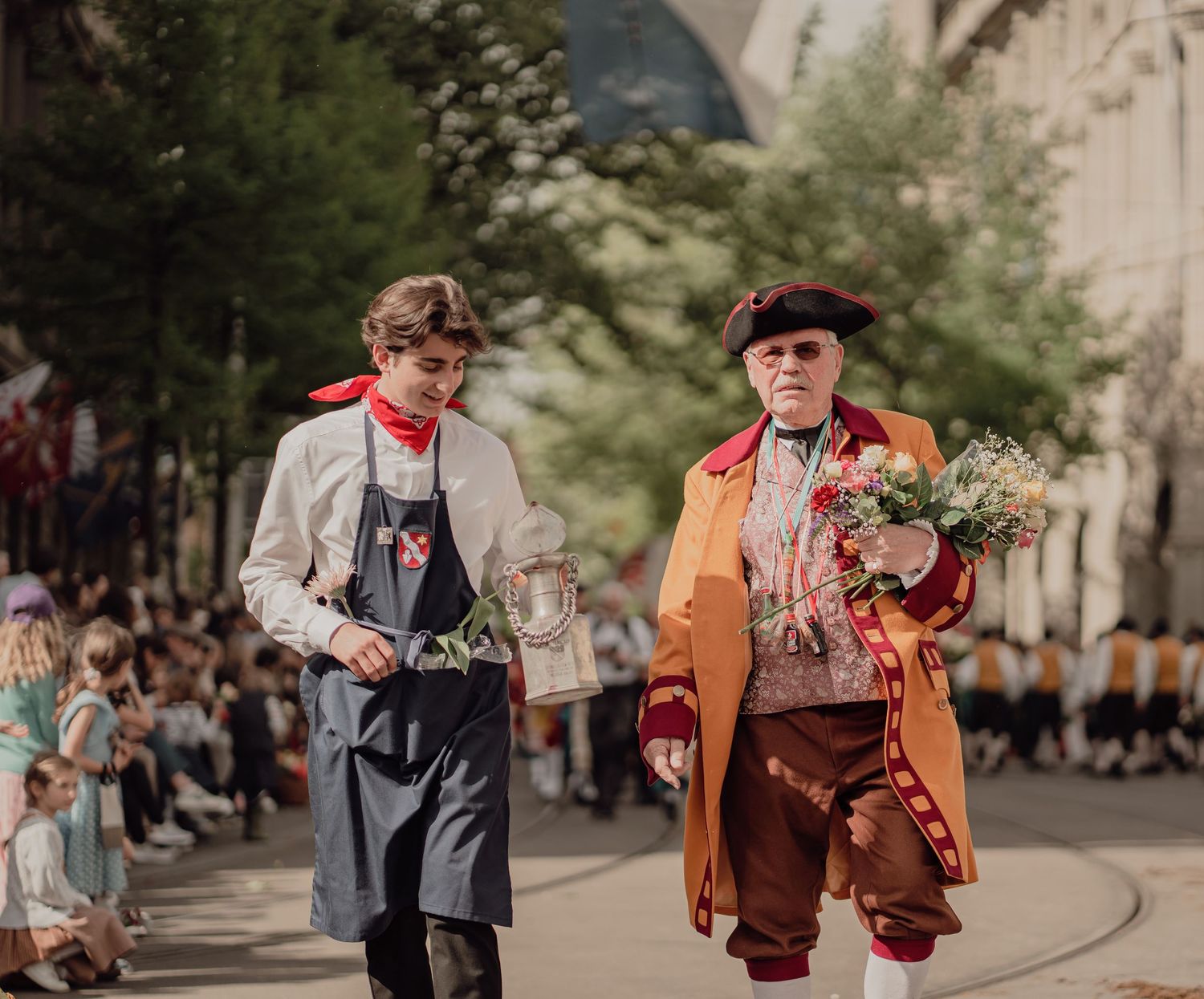 Two men in 18th-century period costumes walk in a sunny street parade carrying flowers.
