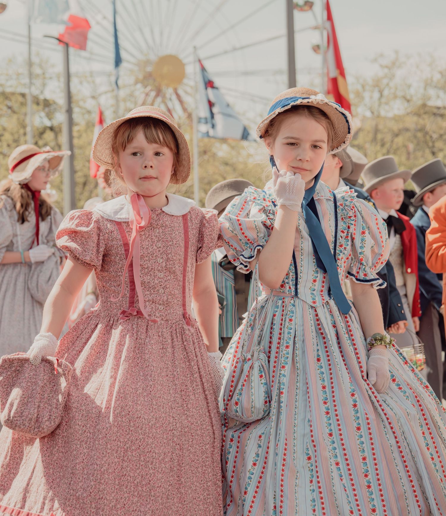 Two young girls in vintage floral dresses and bonnets pose together at a traditional cultural parade.