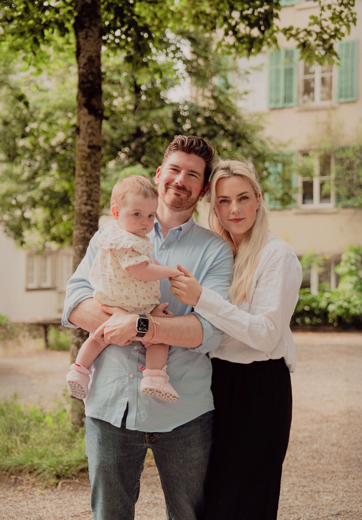Smiling family of three posing together outdoors near green trees in a European garden setting.