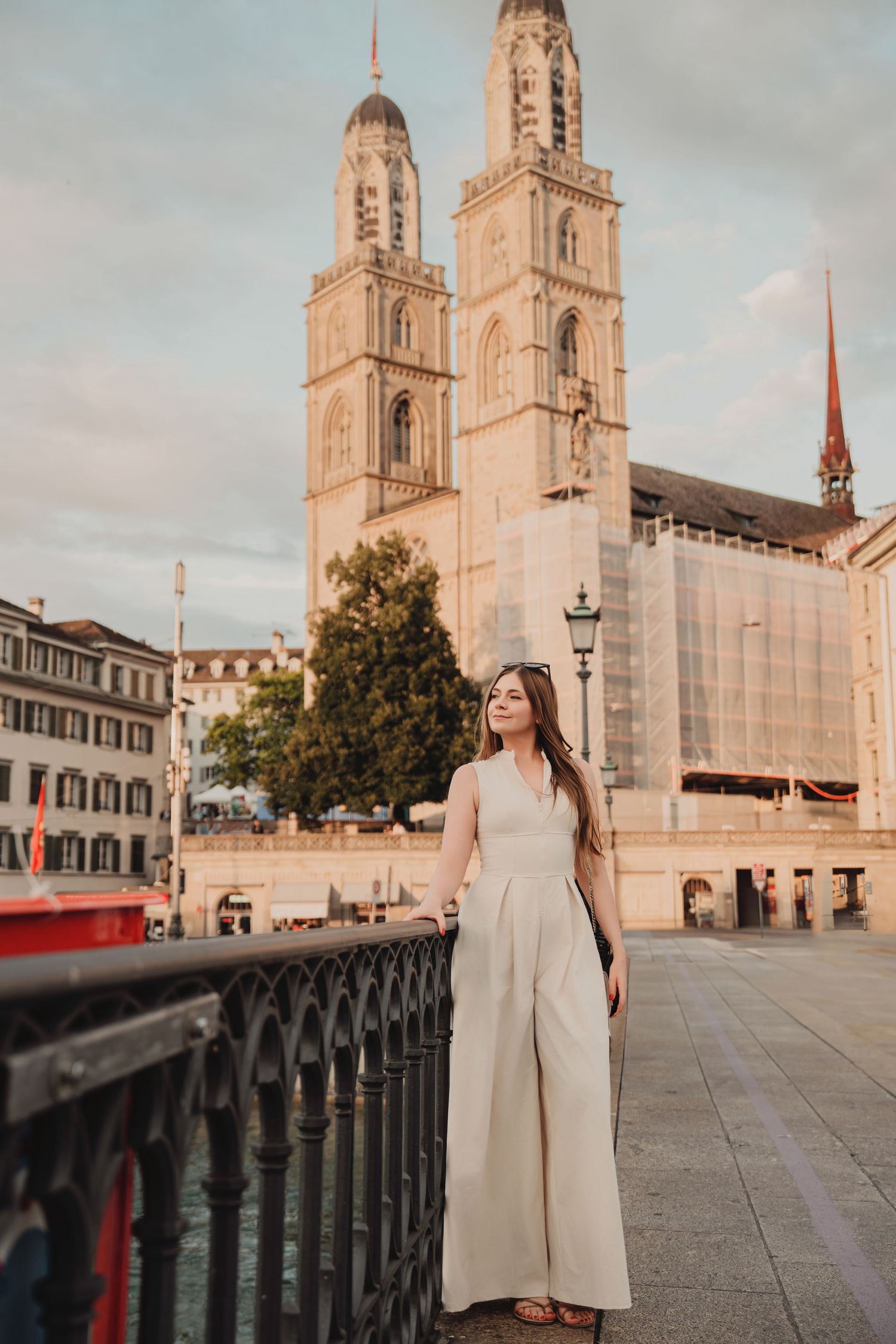 Elegant woman in cream jumpsuit stands near iron railing with Grossmunster Cathedral at golden hour, Zurich.