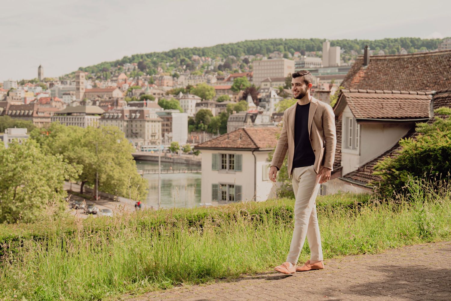 Stylish man in beige blazer walks on sunny Zurich hillside path with historic city buildings in background.