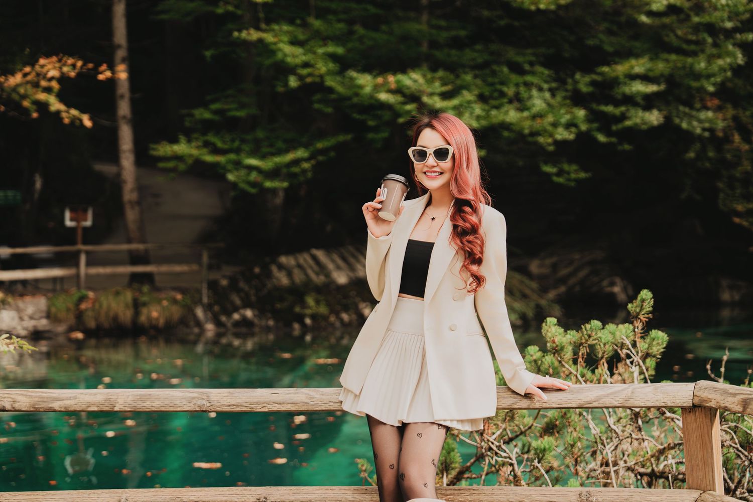 Smiling woman in white blazer with coffee cup posing by turquoise lake in autumn park.