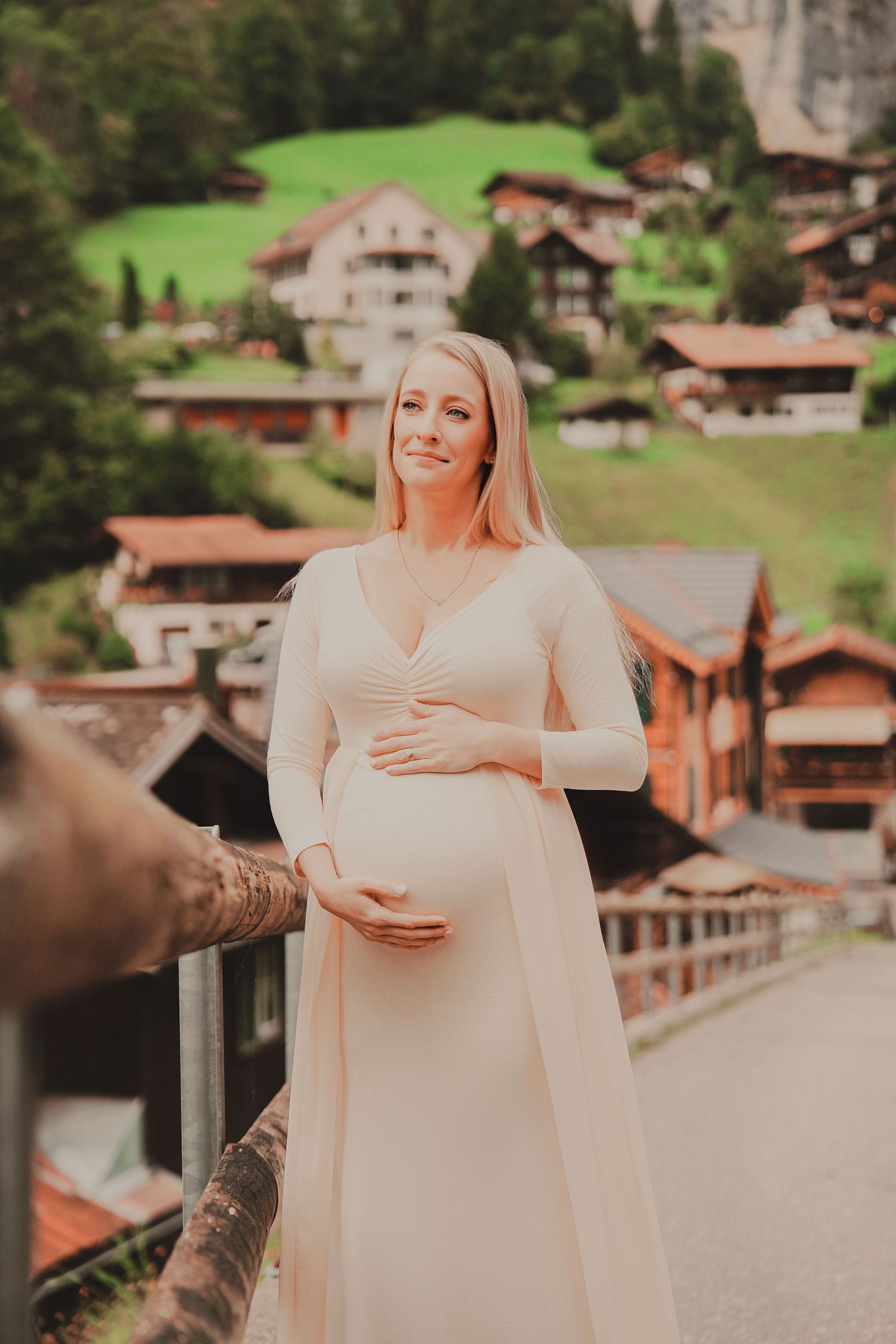 Smiling pregnant woman in cream dress cradling baby bump with charming Swiss alpine village in background.