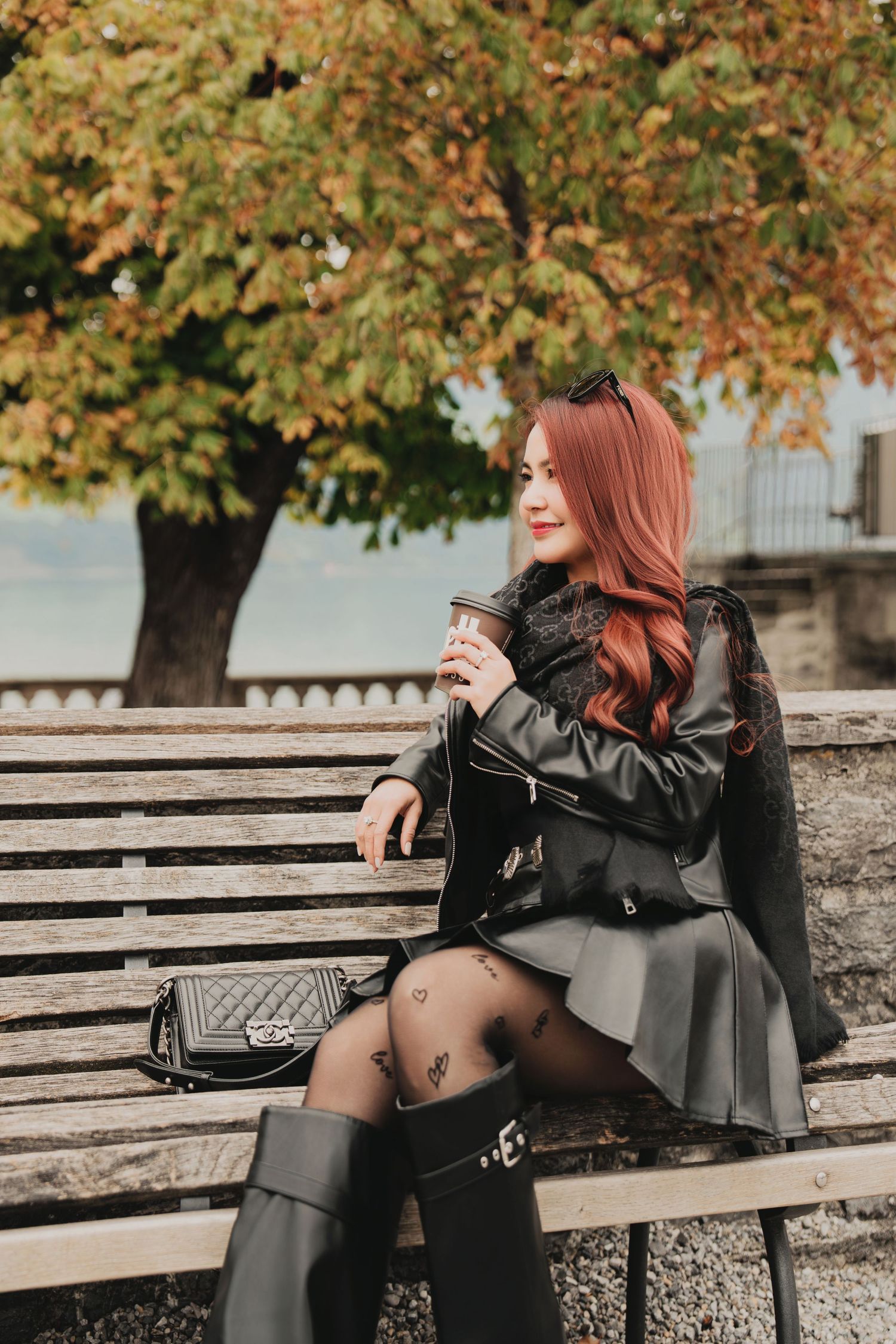 Fashionable red-haired woman in black leather jacket and mini skirt sits on bench with Chanel bag in autumn park.